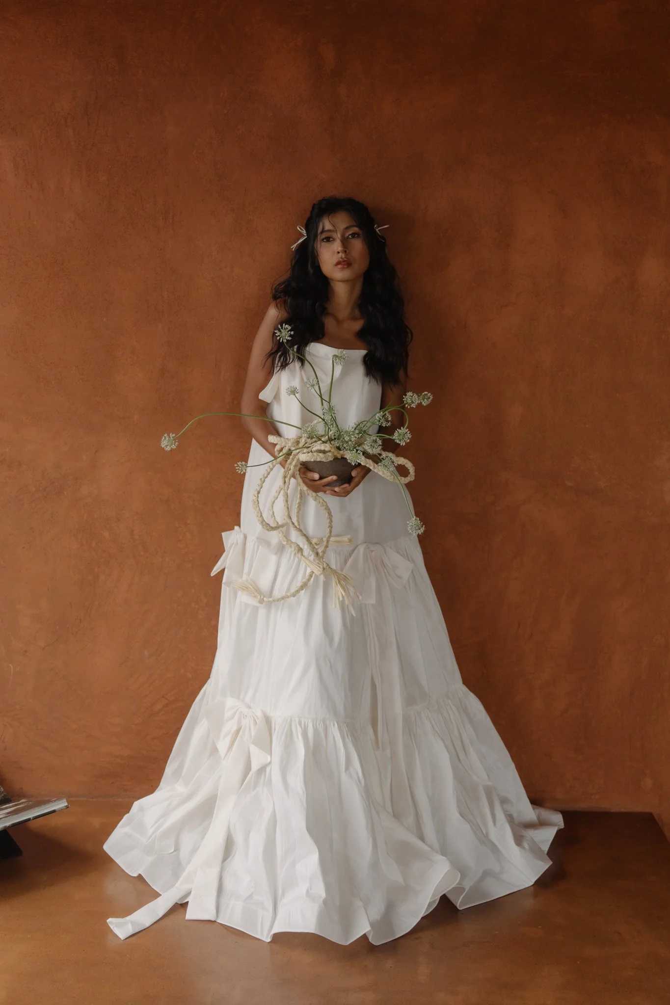 A woman with long wavy black hair, wearing a white dress with ribbons, stands against a brown textured wall, holding a bowl with white flowers and green stems.