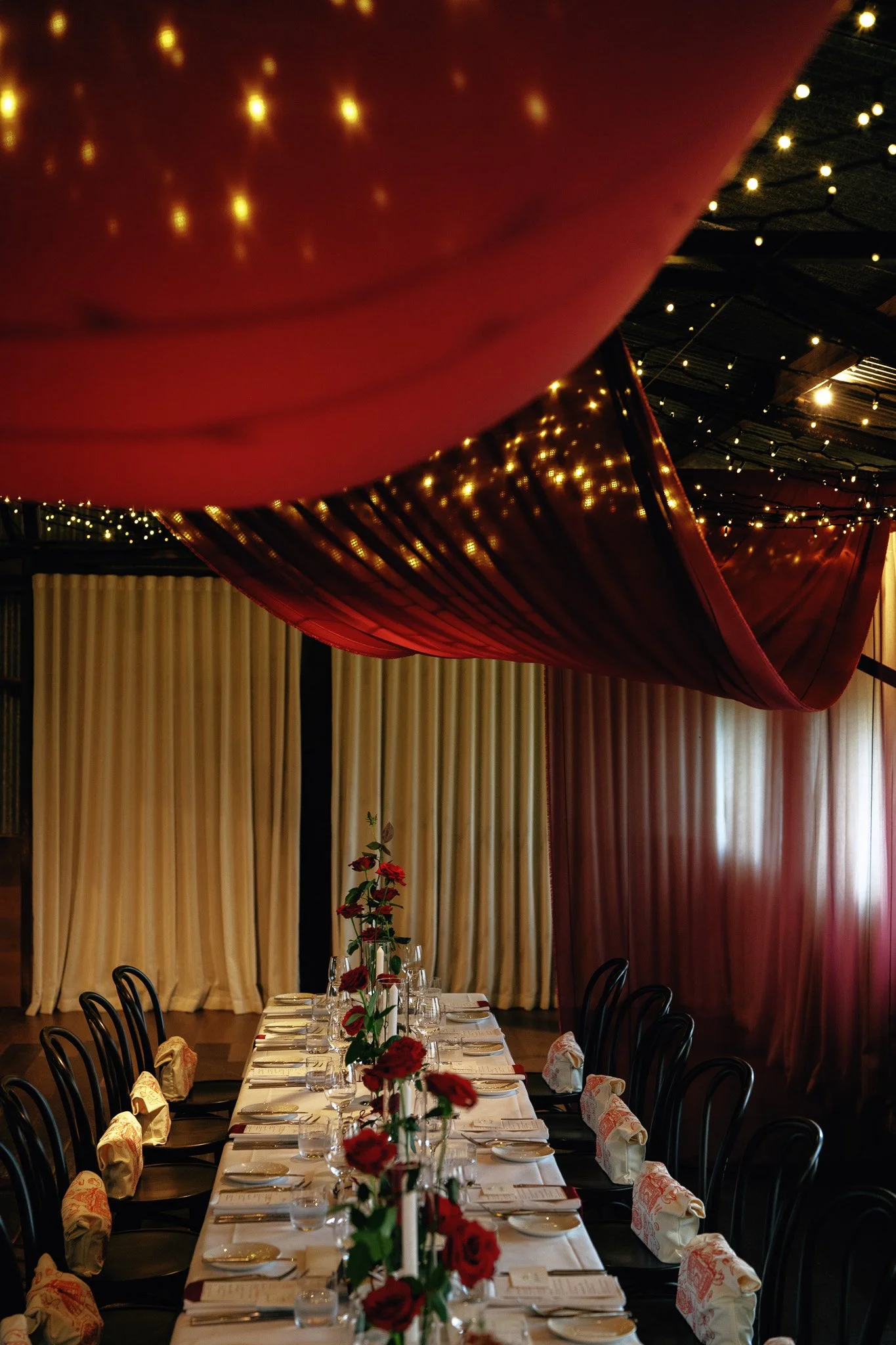 Formal dining setup with a long table adorned with a floral centerpiece, surrounded by black chairs, with cream-colored curtains and decorative drapes and string lights hanging from the ceiling.