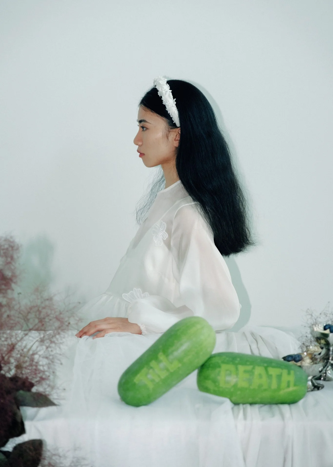 A woman with long black hair wearing a white dress and a white headband, seated with hands resting on her lap against a plain white background.
