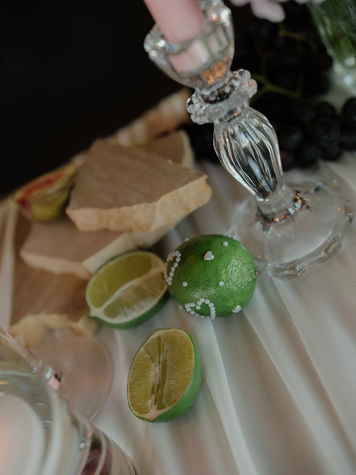 Close-up of a table setting with sliced limes, a decorated lime, a glass decanter, and rocks of ginger and black grapes in the background.