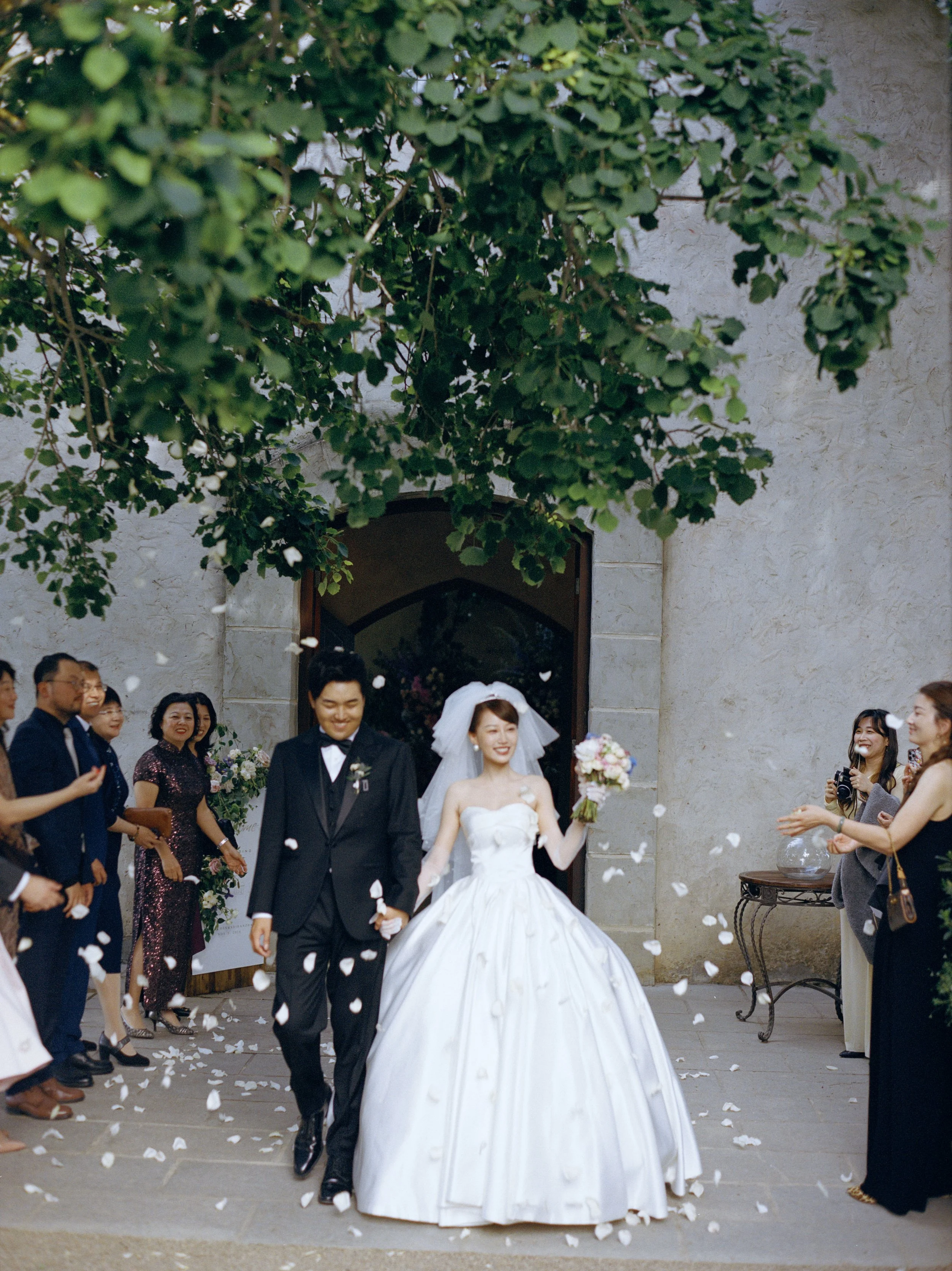 Newlywed couple walking out of a building, surrounded by friends and family celebrating, with flower petals scattered on the ground.