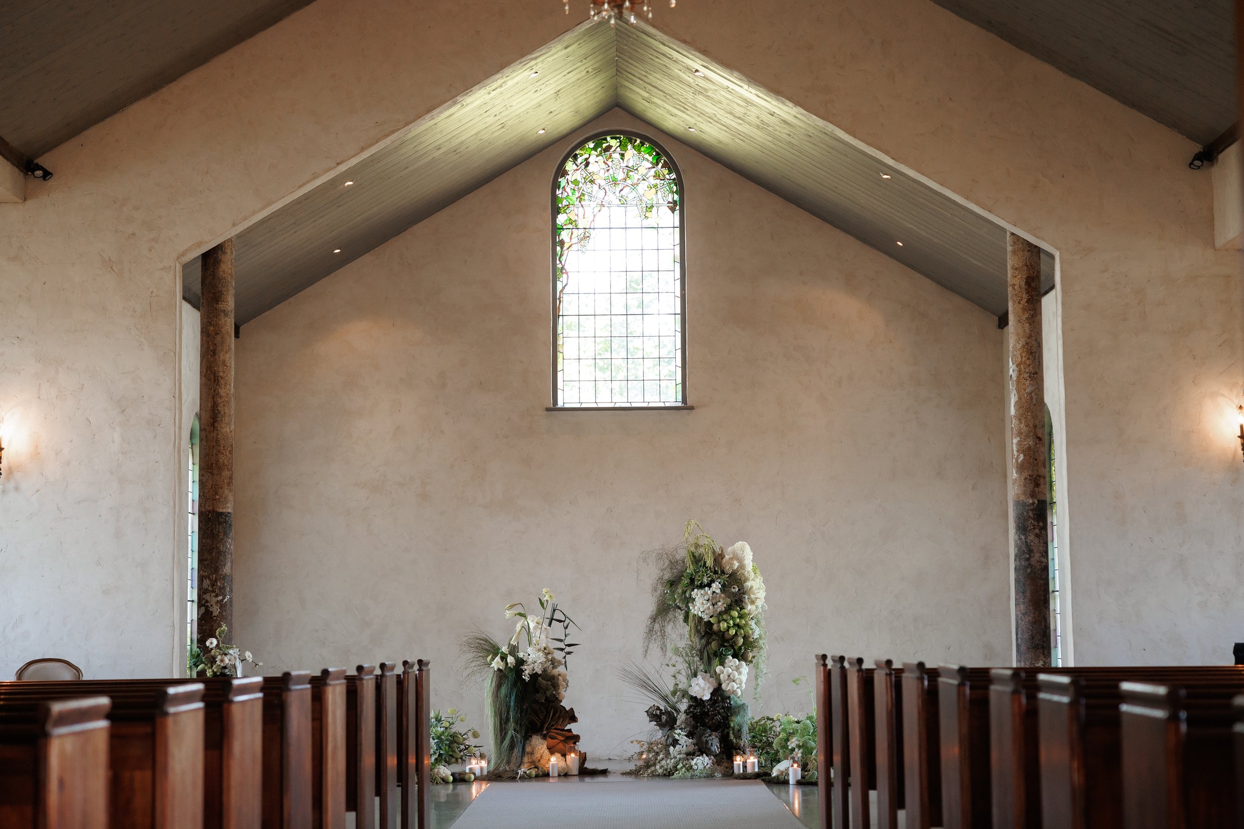 The interior of a church with a high vaulted ceiling and stained glass window at the altar, decorated with flowers and surrounded by candles.
