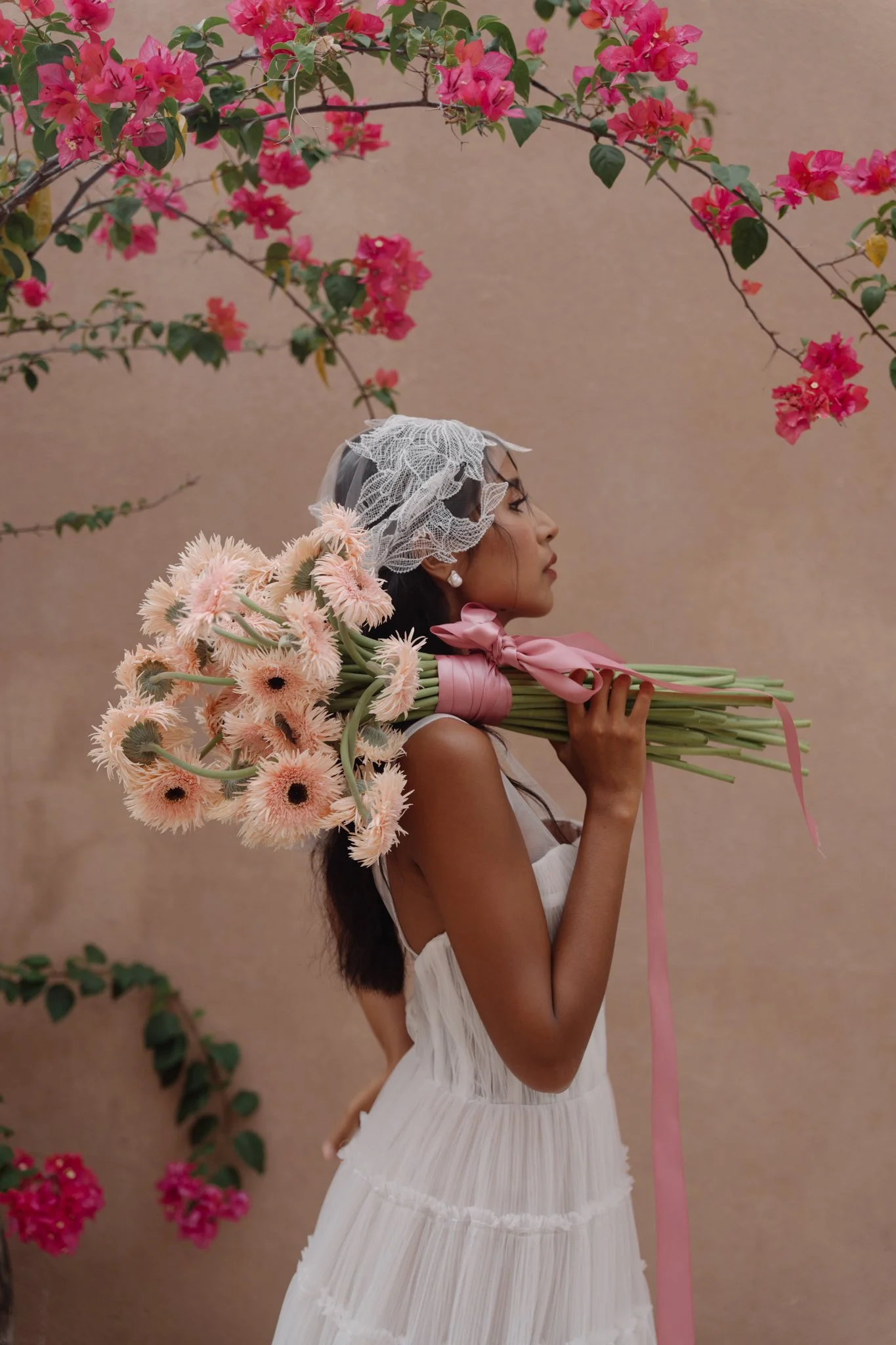 A woman in a white dress holding a large bouquet of pink flowers, standing against a beige wall with pink bougainvillea flowers overhead.