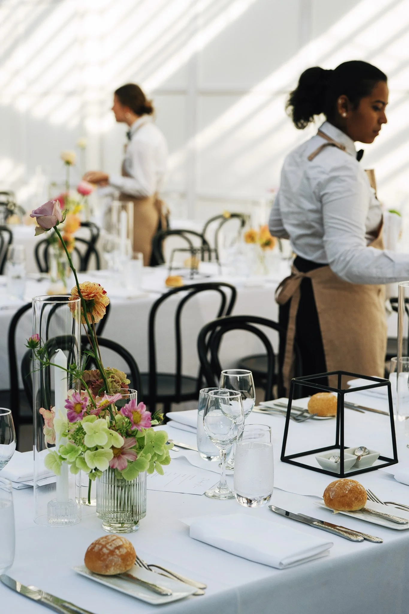 A decorated event table with flower arrangements, wine glasses, bread rolls, and place settings, with two women in uniforms serving in the background.