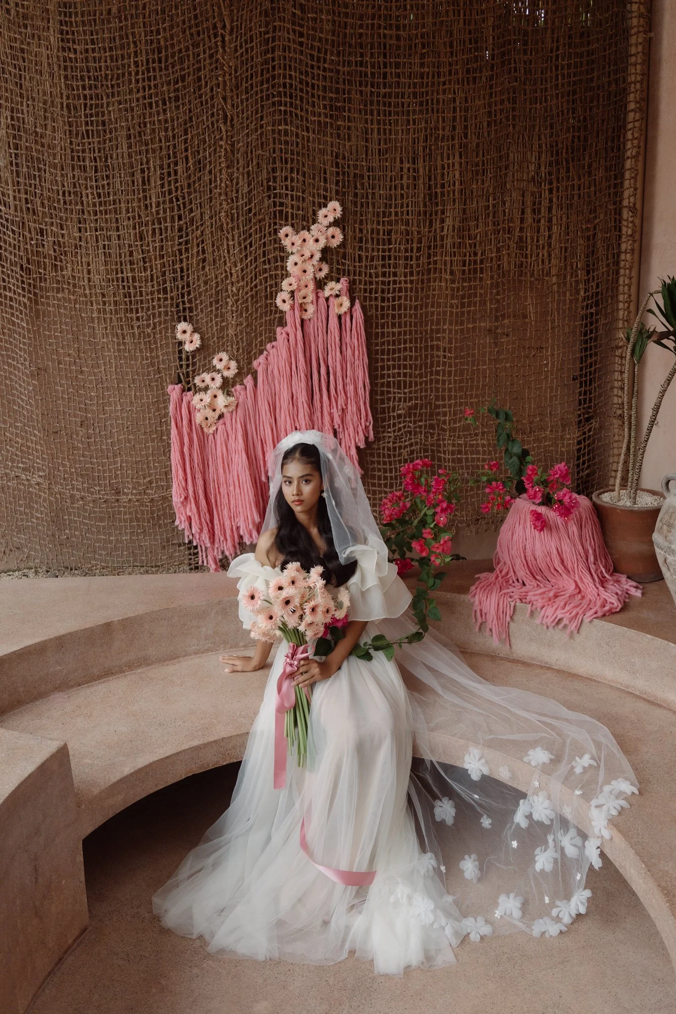 A woman in a white dress and veil sitting on a pink circular stage, holding a bouquet of pink flowers with a pink ribbon. Behind her is a backdrop of brown textured material with pink flowers and pink yarn decorations.