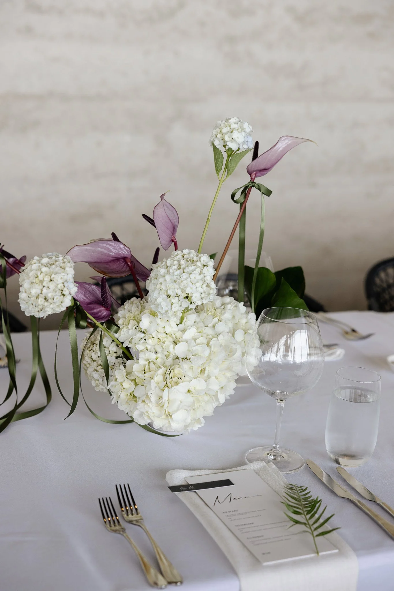 Elegant table setting with floral centerpiece including white hydrangeas and purple calla lilies, wine glass, water glass, silverware, menu card, and a fern leaf on a white tablecloth.
