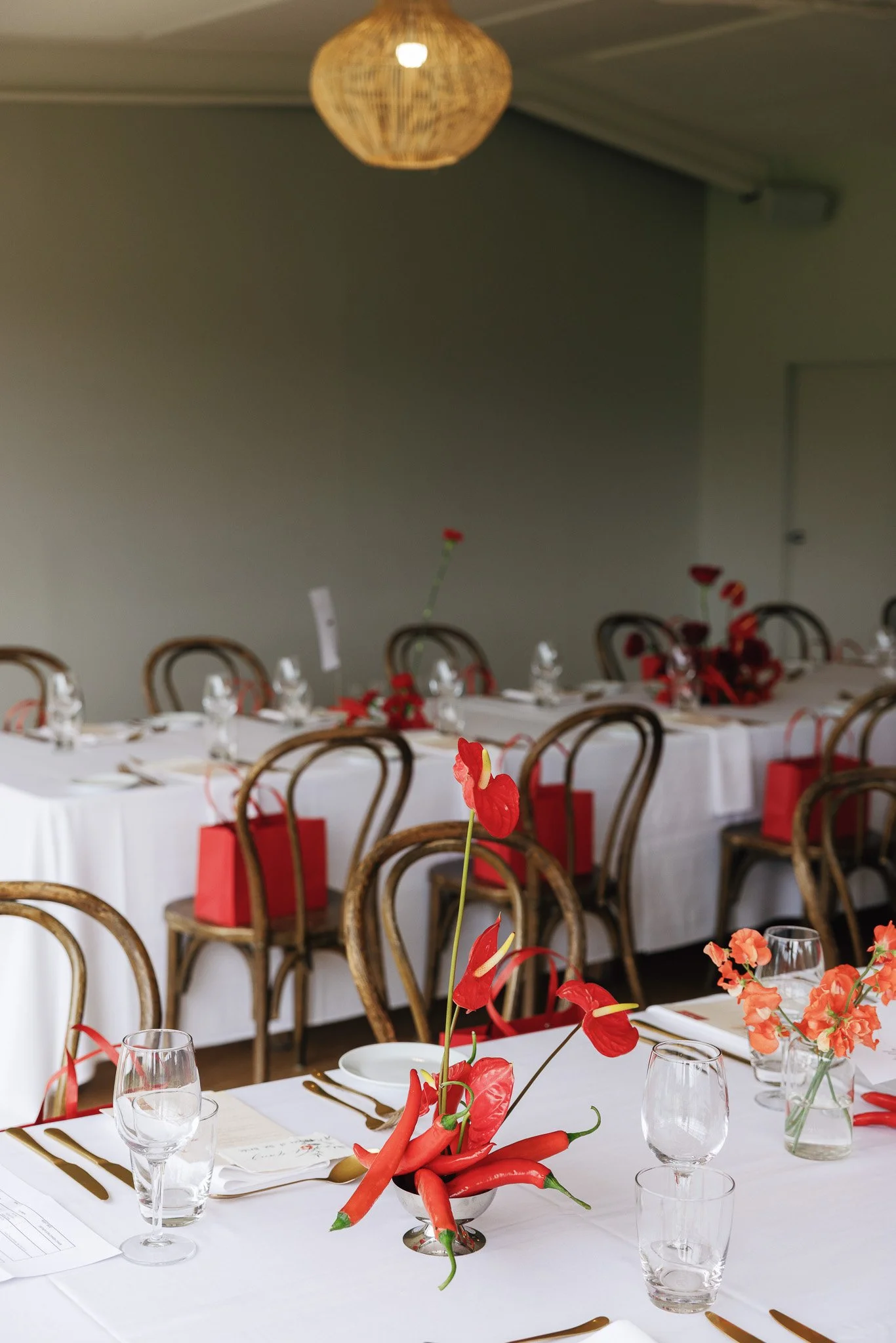 A dining table decorated for a special occasion with white tablecloths, floral centerpieces with red and orange flowers, glasses, and gold-rimmed plates. The background features multiple chairs and a grey wall with a wicker pendant lamp hanging from 