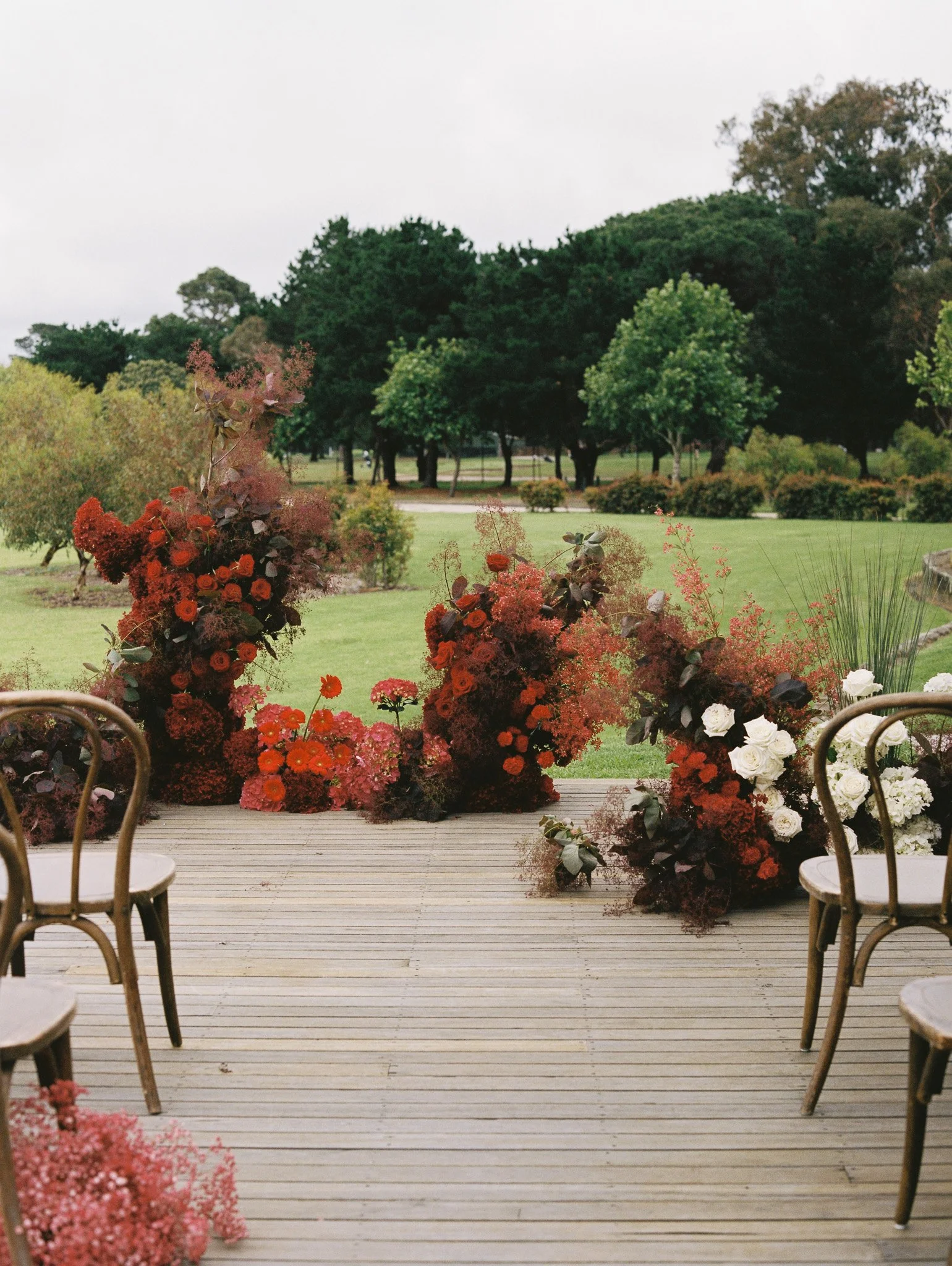Outdoor garden scene with a wooden deck, chairs, and floral arrangements, overlooking a grassy area with trees.
