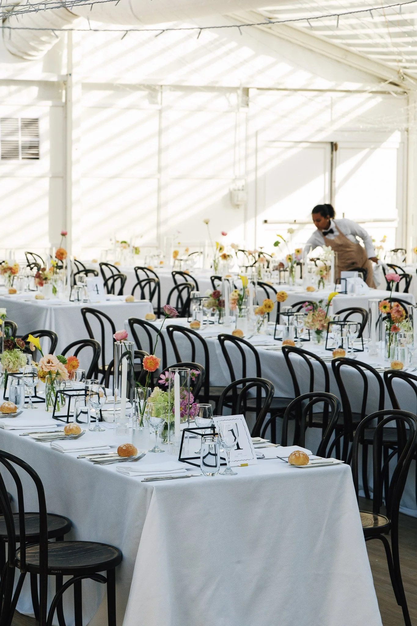 Elegant banquet hall with round tables covered in white tablecloths, decorated with colorful flower centerpieces, glassware, and place settings, set for a formal event or wedding reception. A server is adjusting a table in the background.