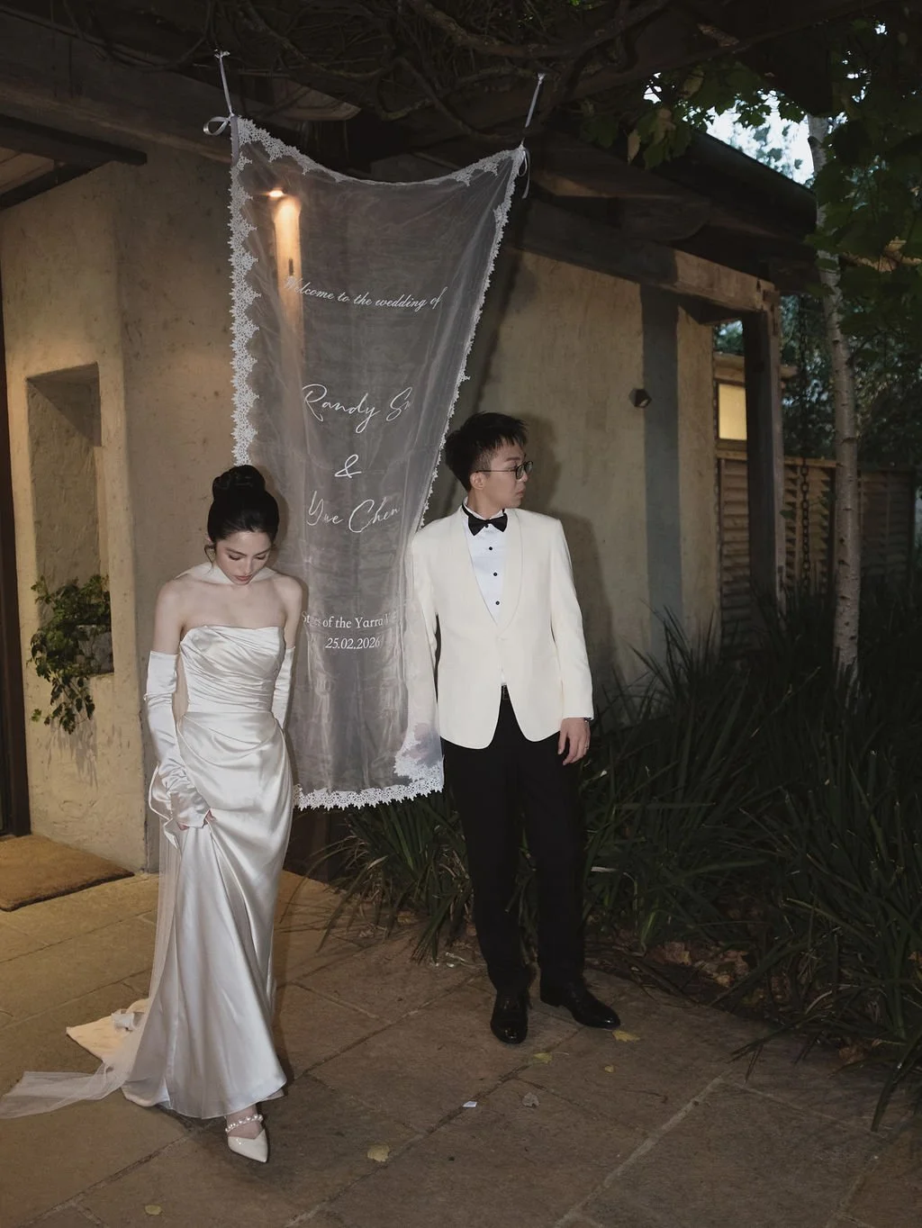 A bride and groom standing side by side outside, dressed in wedding attire, in front of a semi-transparent wedding sign with cursive writing.