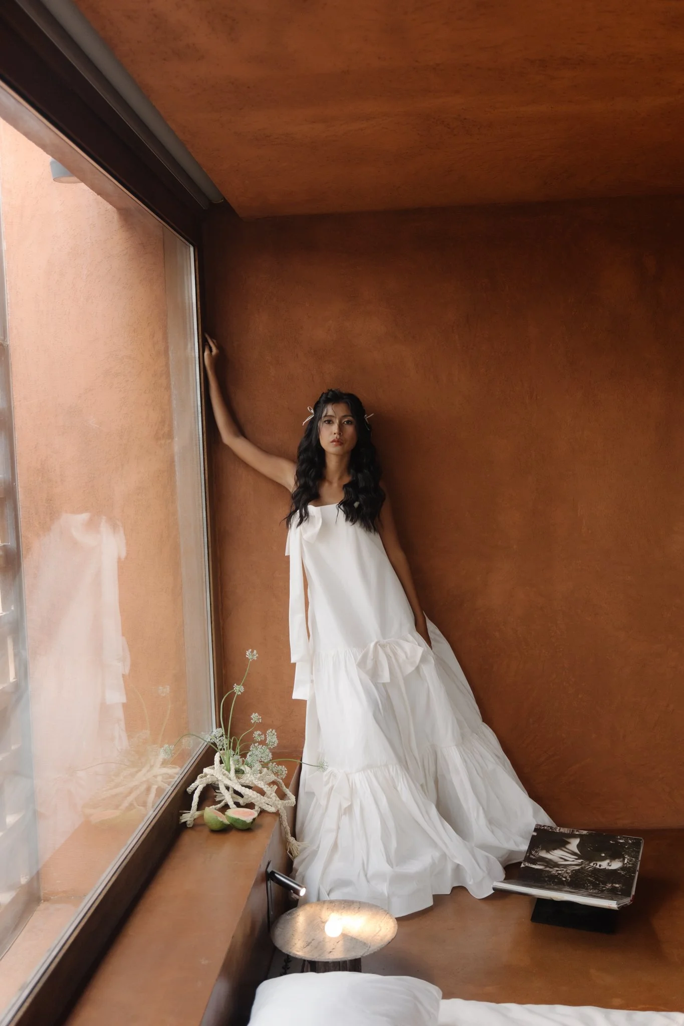 A woman in a white dress with long dark hair standing in a corner of a room with wooden walls and a large window, holding the wall with one hand. A small table at the window holds a vase with white flowers and some plush toys. There is an open book a