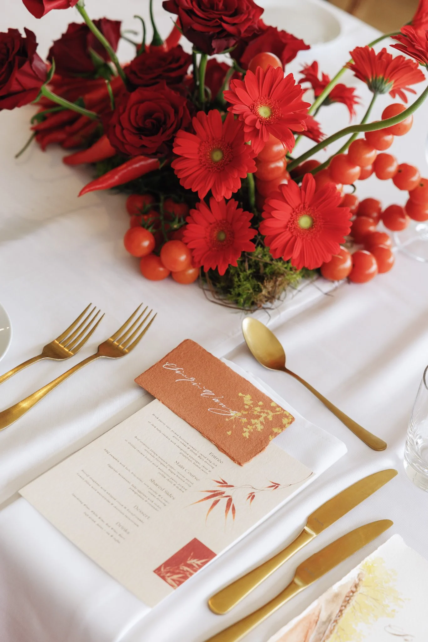 A centerpiece of red flowers, including roses and daisies, with green stems, arranged on a white tablecloth. Gold cutlery and a printed menu are also visible on the table.