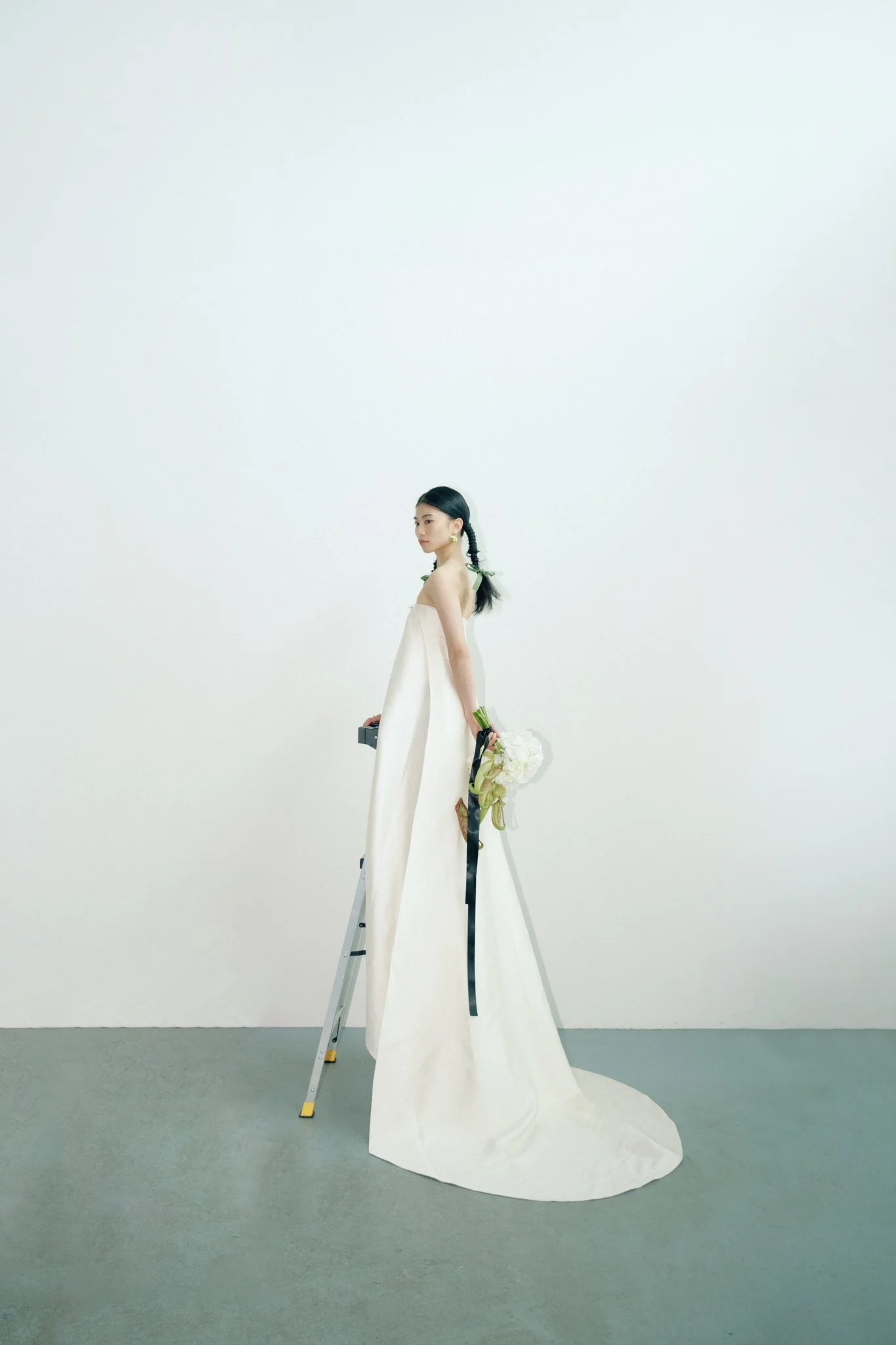 A woman in a white dress stands on a ladder, holding flowers and a box, against a blank white wall.