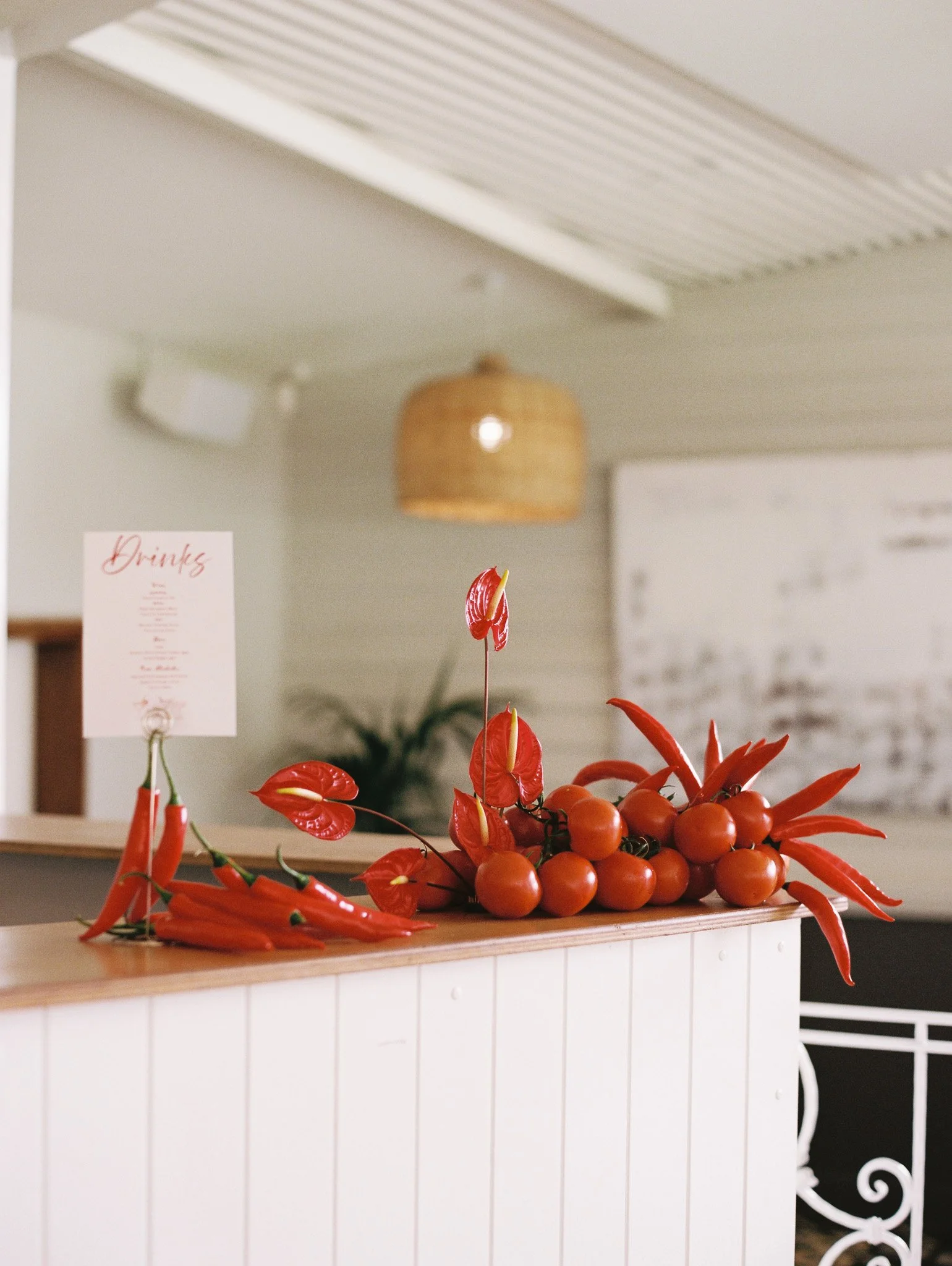 Arrangement of red anthurium flowers and small orange tomatoes on a wooden surface in front of a white wall.