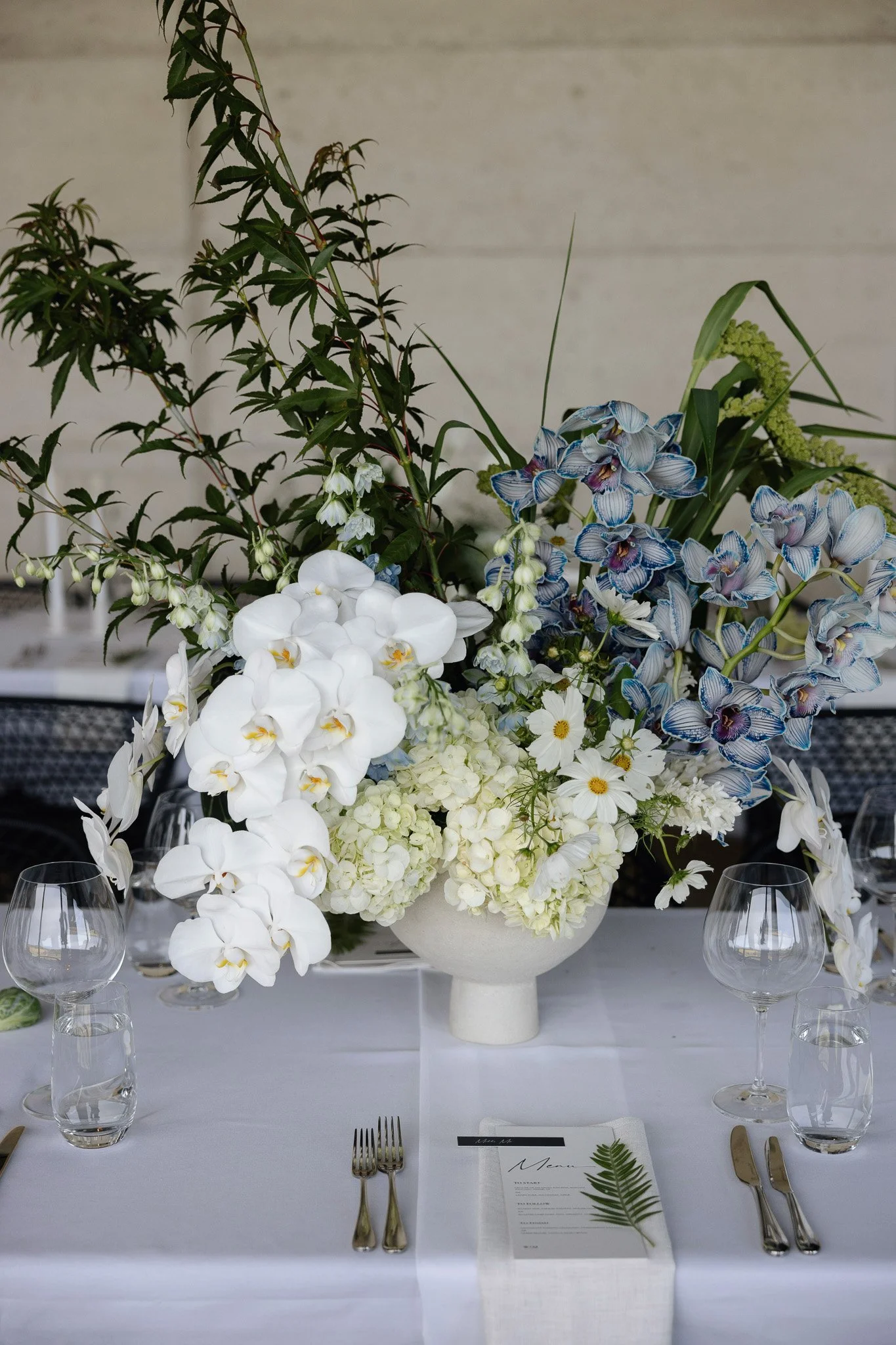 A formal dining table with a white tablecloth featuring a large floral centerpiece in a white vase. The arrangement includes white orchids, blue and white striped orchids, and other white flowers, accented with greenery. The table is set with clear w