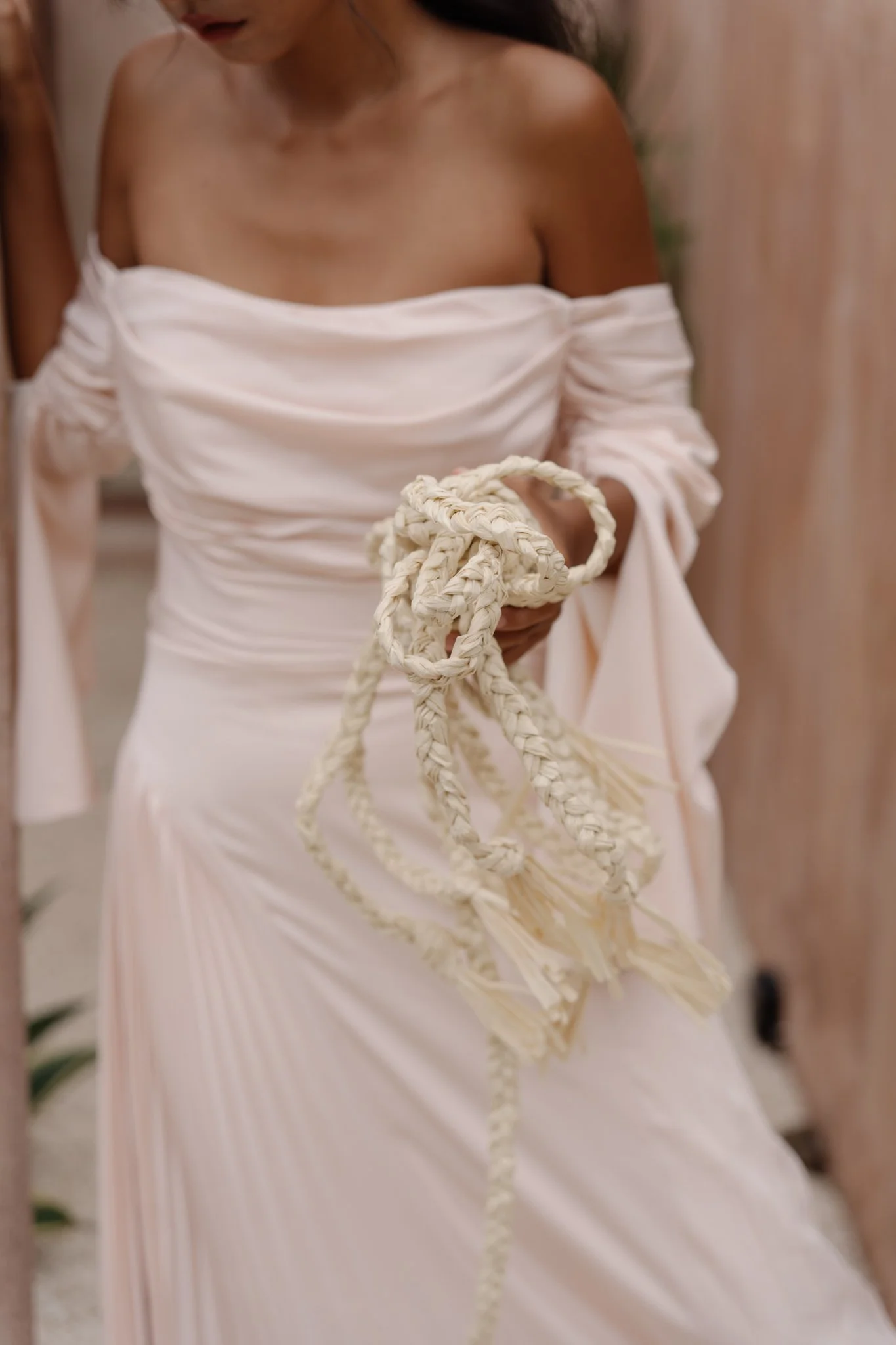 A woman dressed in an off-shoulder white gown holding a cream-colored braided rope, standing indoors with a blurred background.