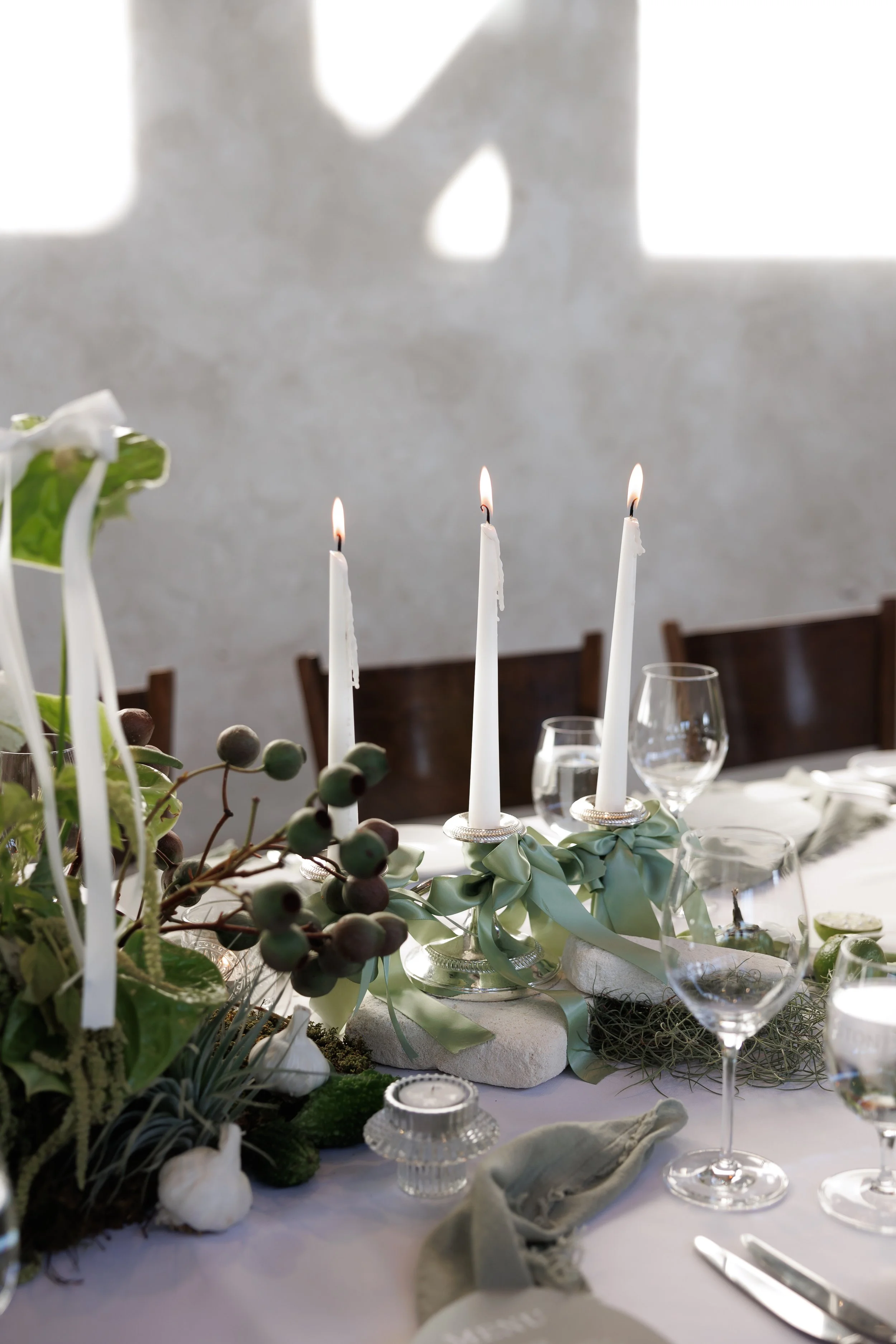 A decorated dining table with white candles in silver holders, floral arrangements with greenery and berries, glassware, and a soft white tablecloth.