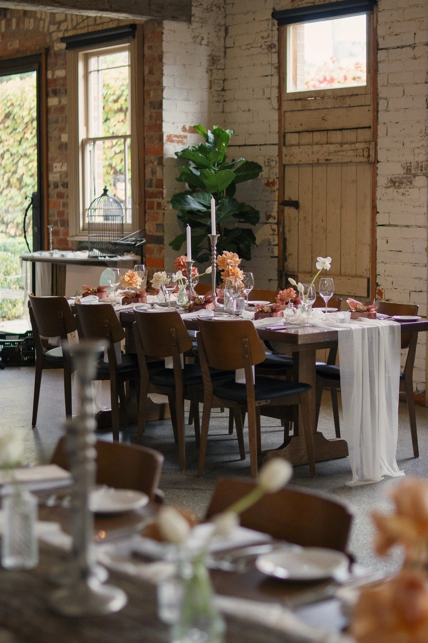A decorated dining table set for a formal event in a rustic indoor space with exposed brick walls and windows.