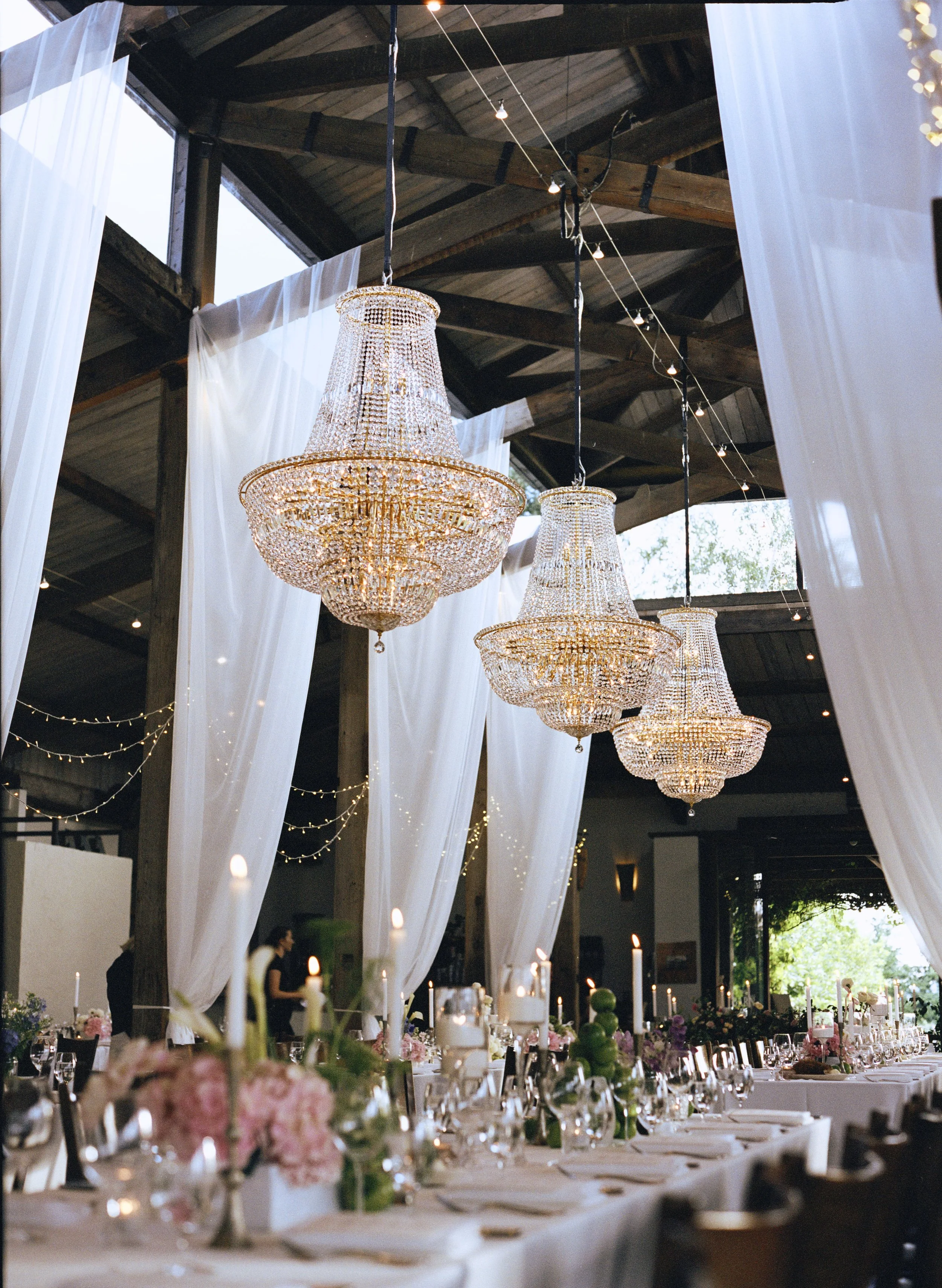 Elegant indoor event space decorated with large crystal chandeliers hanging from a high wooden ceiling. White drapes are hung around the room, illuminated by string lights. A long table is set for a celebration, with flowers, candles, glassware, and 
