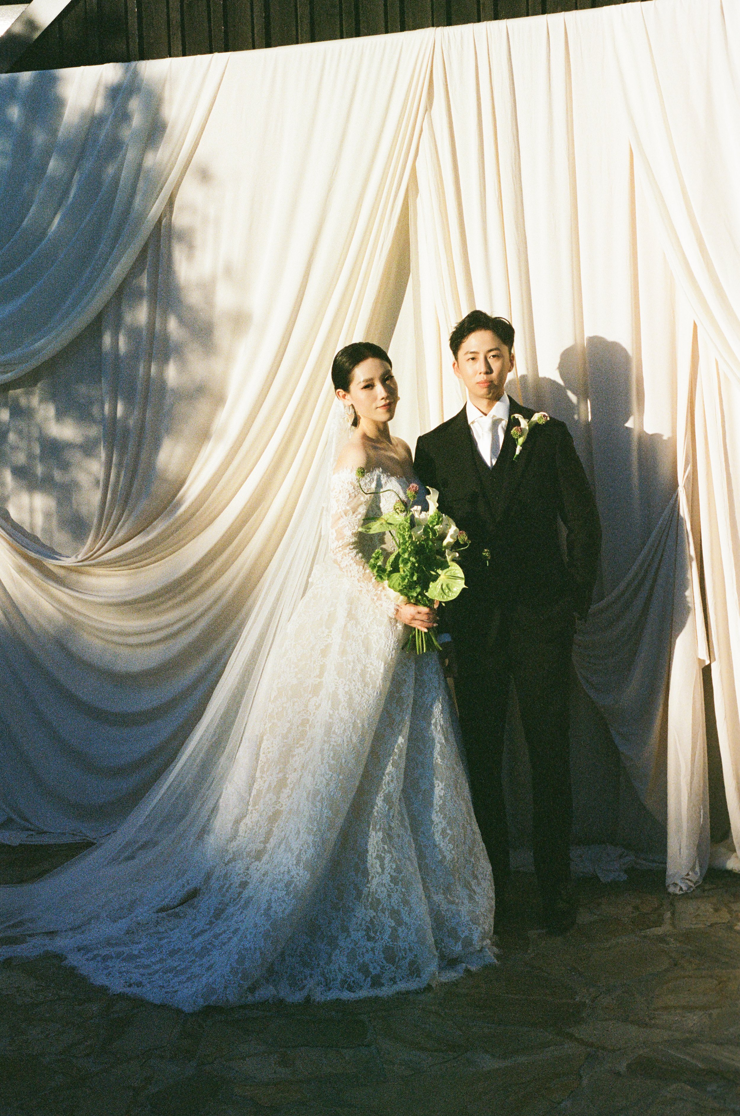 A bride and groom standing together in front of cream-colored curtains, with the bride holding a bouquet of flowers.