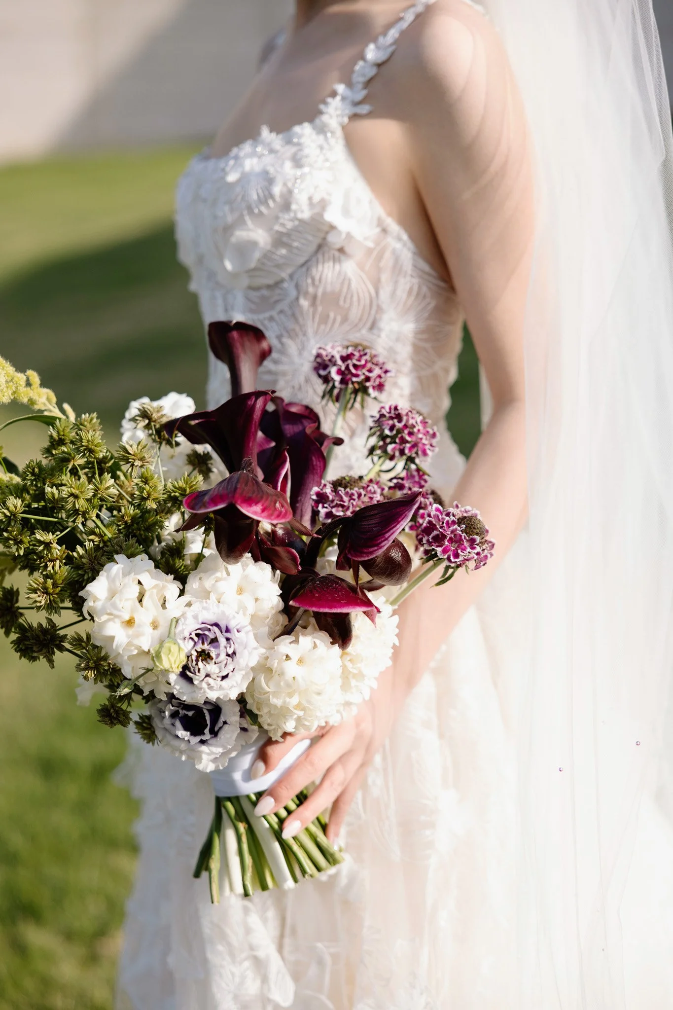 A woman in a wedding dress holding a bouquet of flowers, standing outdoors on a grassy area.