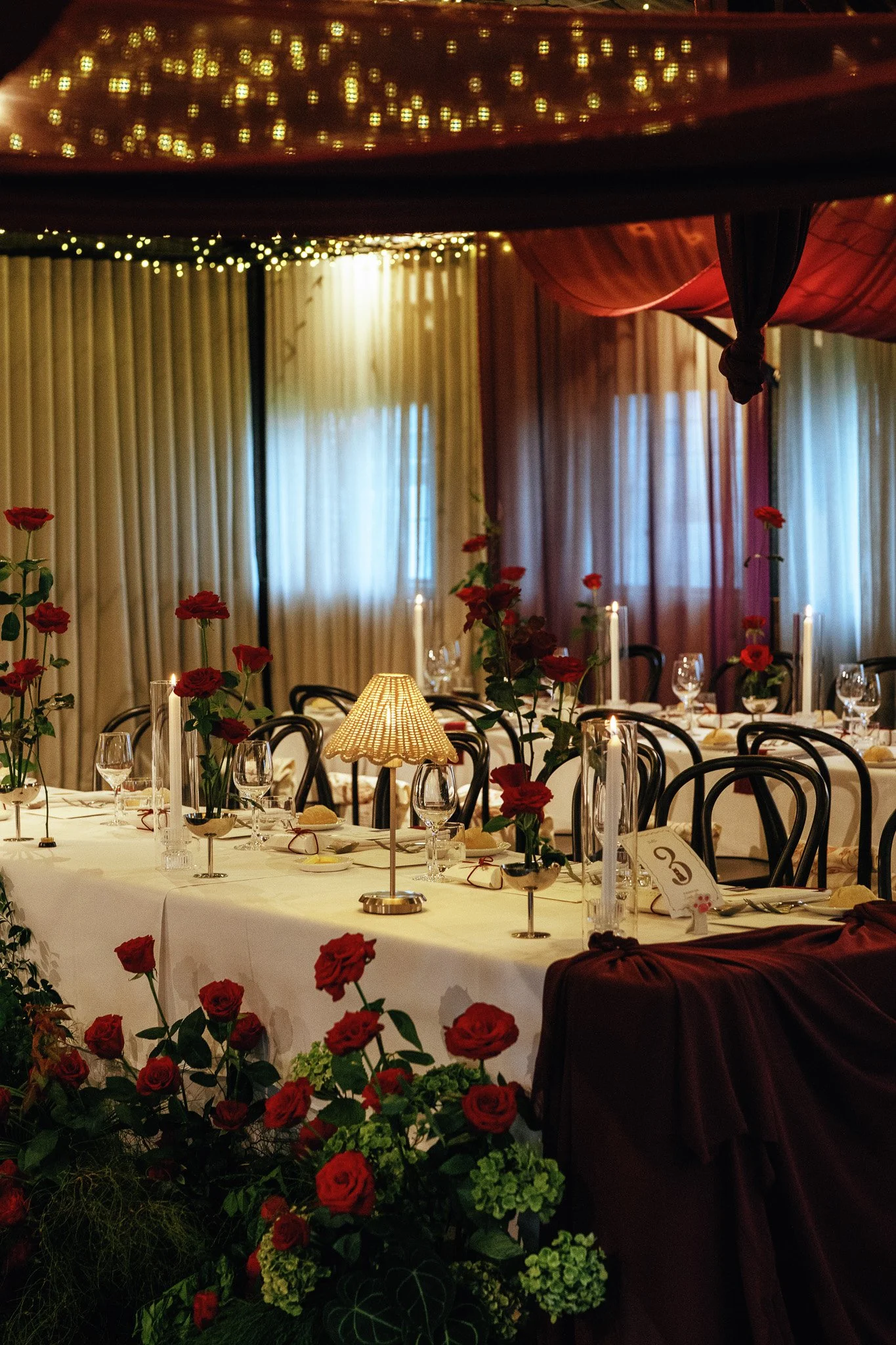 A decorated banquet table with red roses, candles, and a small lamp in a warmly lit dining room, possibly for a special occasion or celebration.