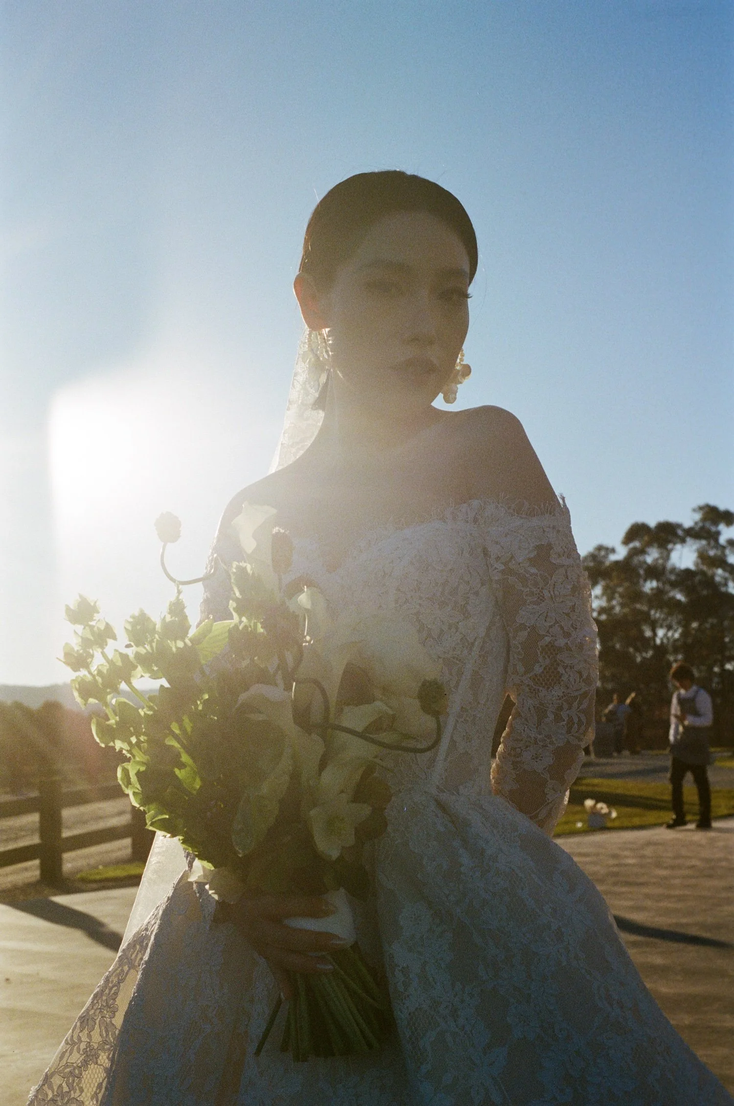 A woman in a white lace dress holding a bouquet of white flowers, standing outdoors with the sun behind her, creating a glowing effect around her silhouette.