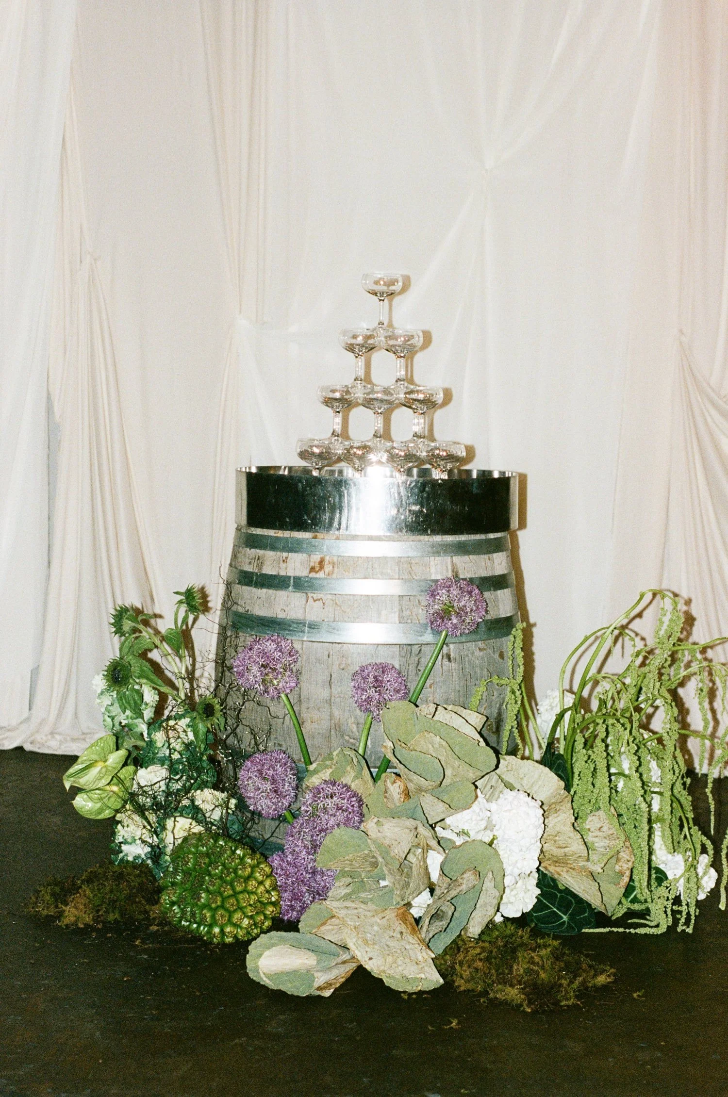 A champagne bucket with a pyramid of empty glasses on top, surrounded by various green and purple plants and flowers, set against a cream-colored draped backdrop.