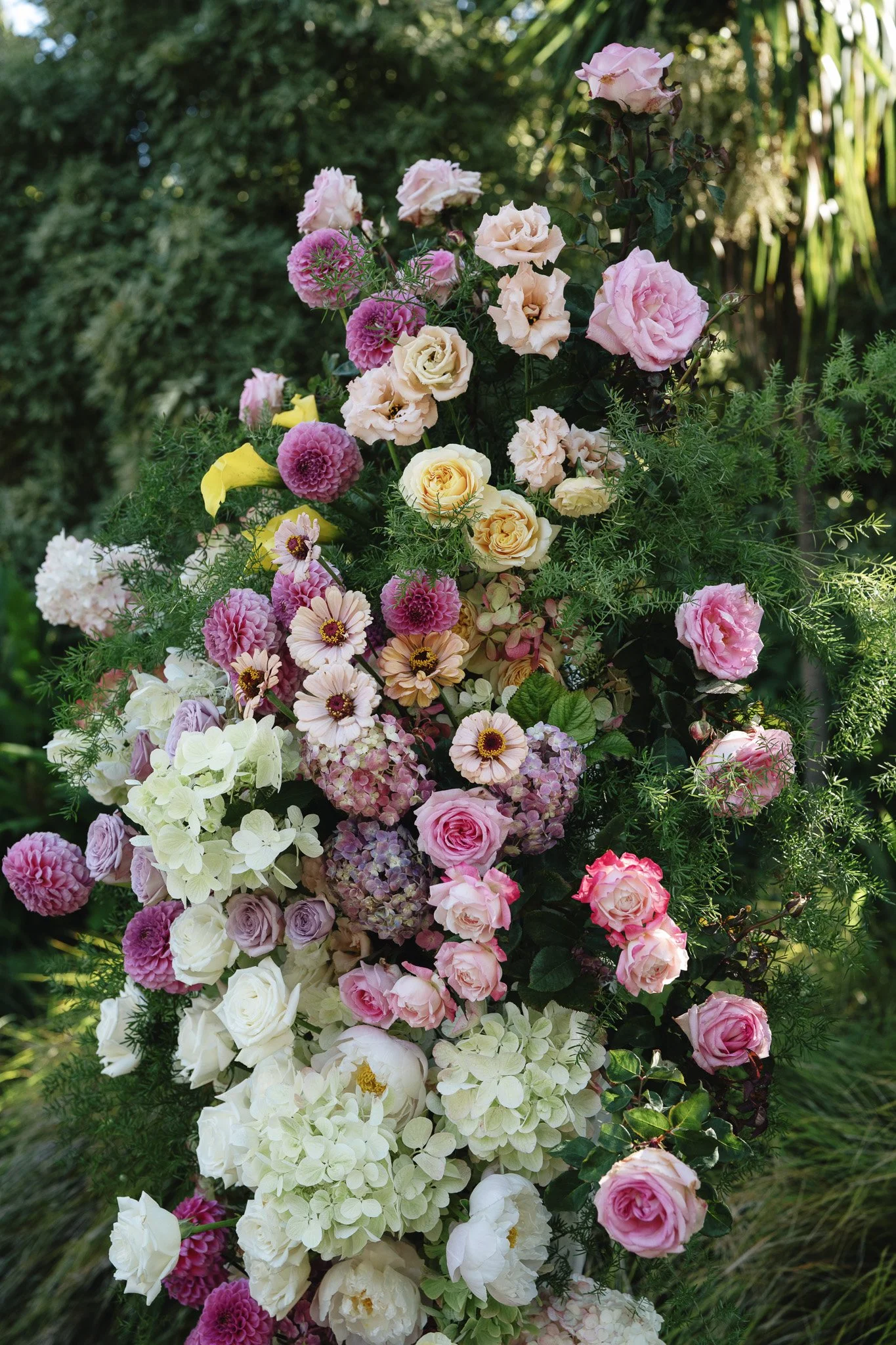 A colorful arrangement of various flowers including roses, hydrangeas, dahlias, and daisies in shades of pink, white, and yellow, set against a green garden background.