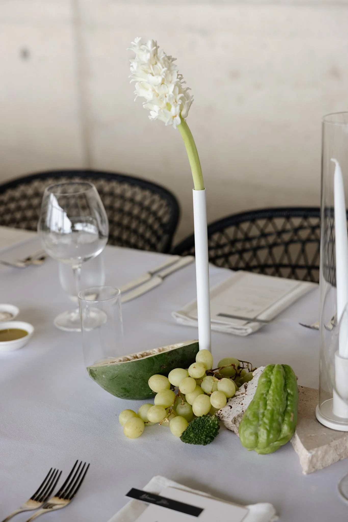 A white table with a centerpiece that includes a white hyacinth flower in a white holder, green grapes, and a piece of white coral, surrounded by several wine glasses and utensils in a well-lit indoor setting.