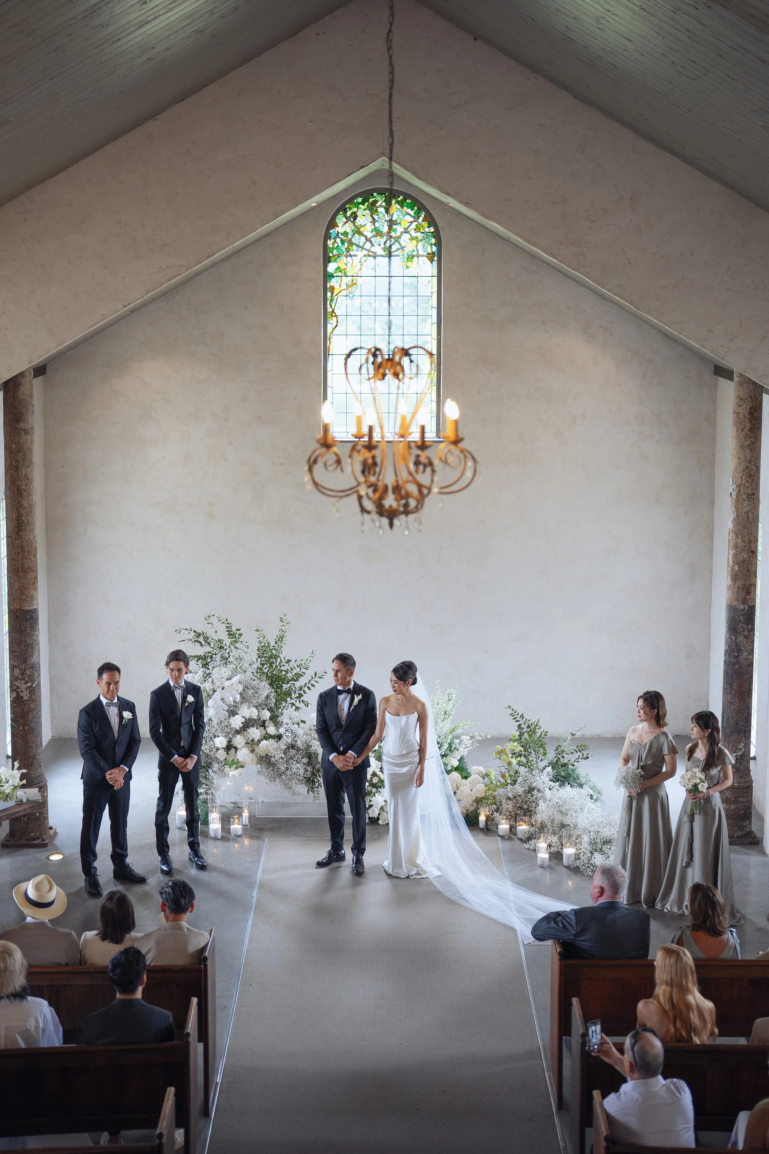 A wedding ceremony taking place in a church with a high peaked ceiling, featuring large stained-glass windows and a chandelier, with the bride and groom standing in front of a floral arrangement with white flowers and greenery, while guests are seate