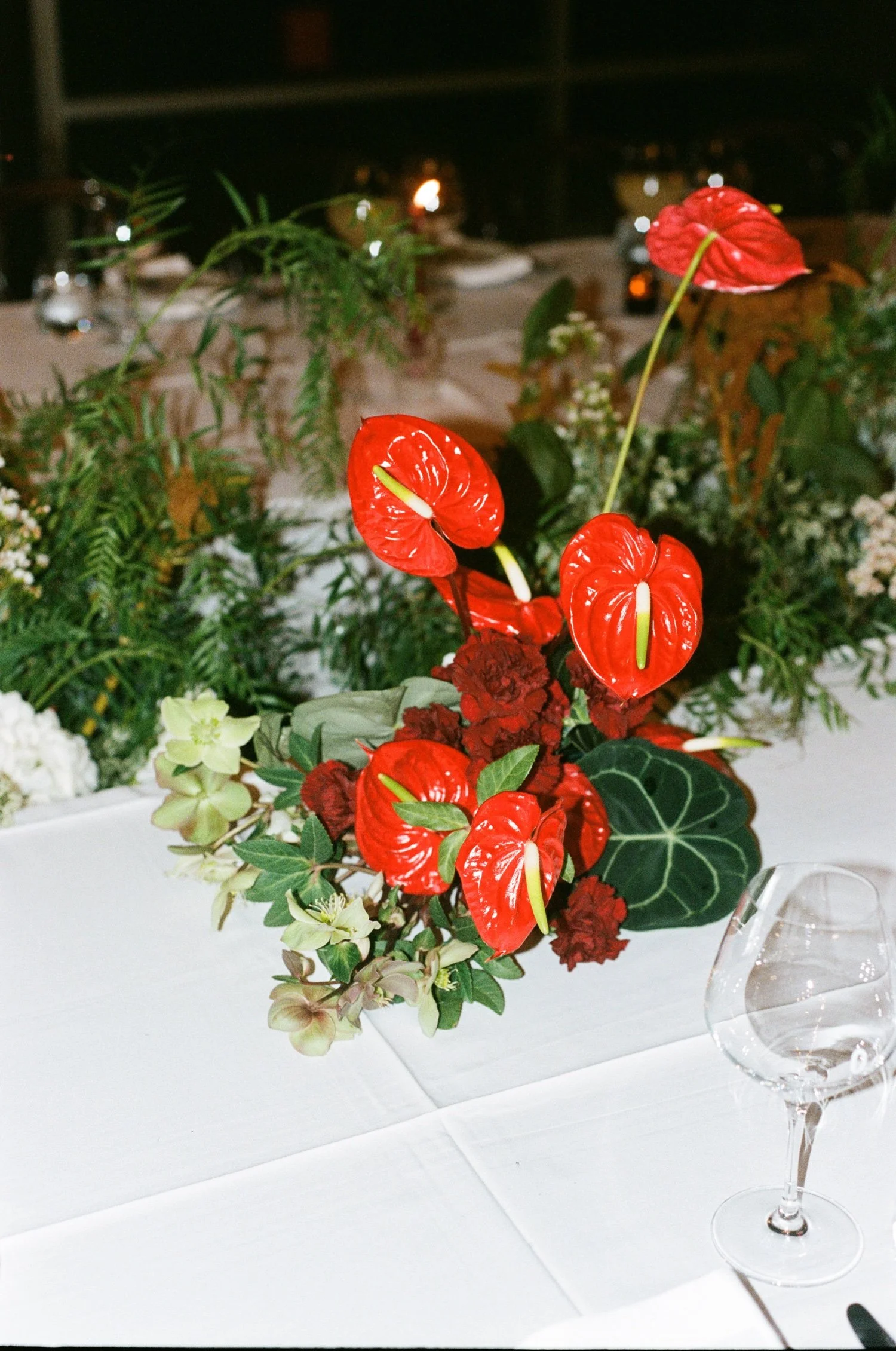 A floral centerpiece with red anthuriums, green leaves, and other flowers on a white table at night, with a wine glass nearby.