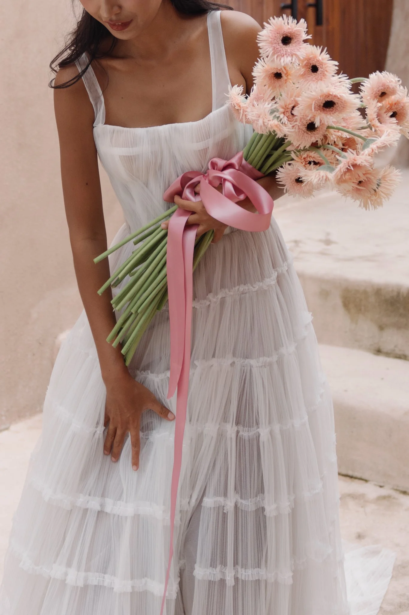 Close-up of a woman in a white pleated dress holding a large bouquet of pink and peach flowers with a pink satin ribbon bow.