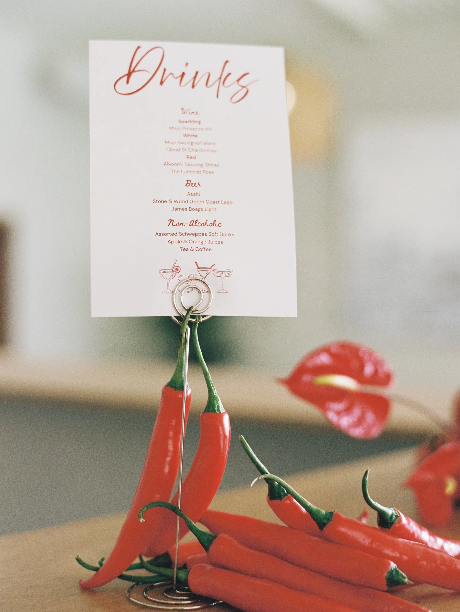 A collection of red, orange, and green chili peppers on a table in front of a white drink menu with red cursive and regular lettering listing wines, beers, and non-alcoholic drinks.