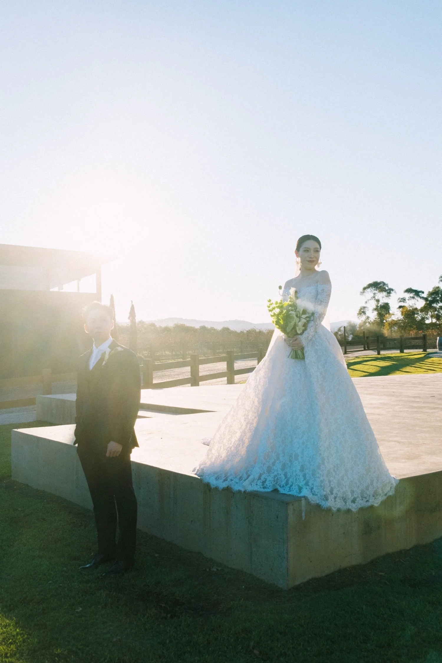 A bride in a wedding dress holding a bouquet, standing on a ledge outdoors during sunset, with a young boy in a suit standing nearby.