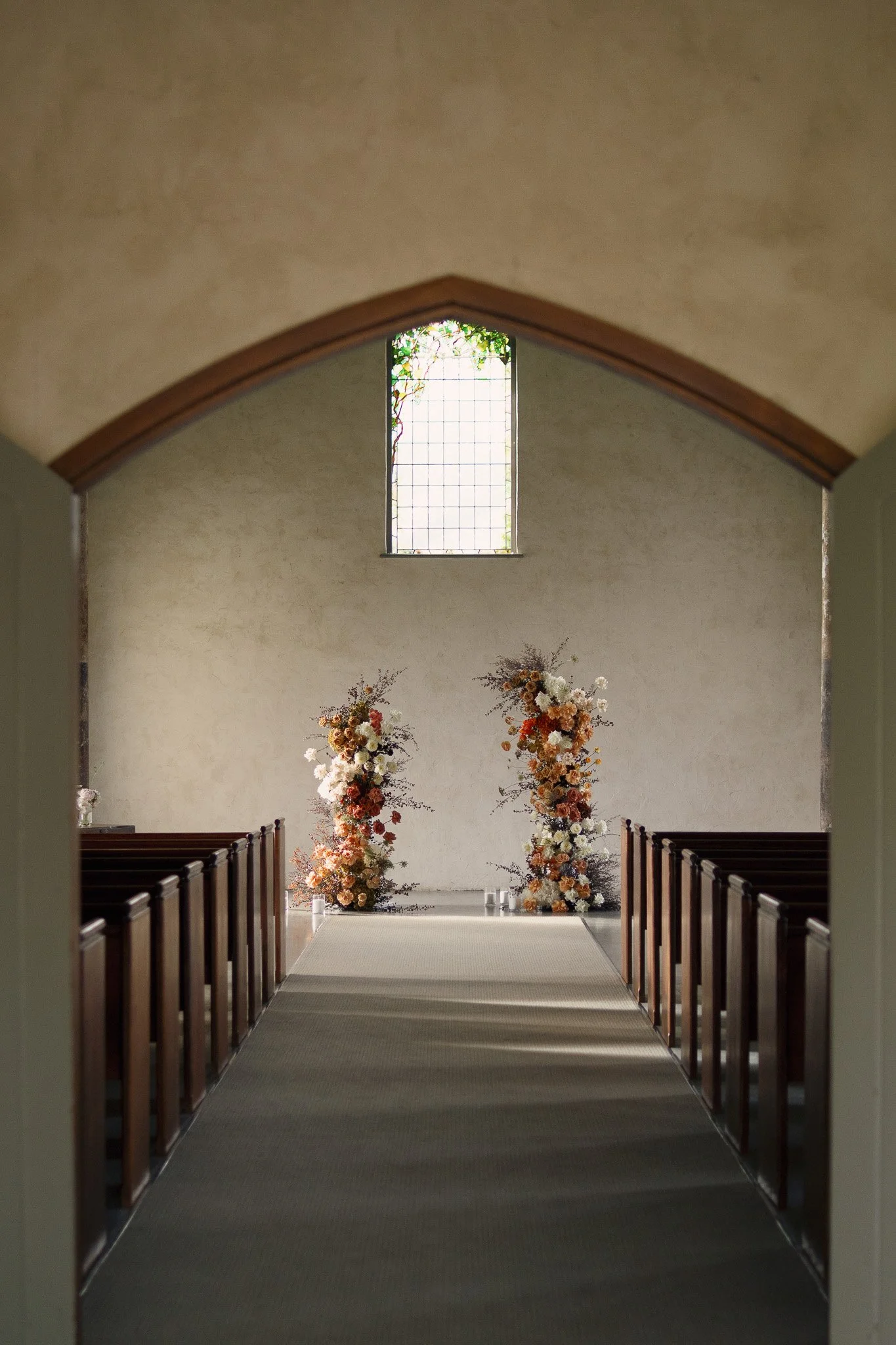 Interior of a church with altar decorated with large floral arrangements and candles, viewed through an arched doorway with wooden pews on either side.