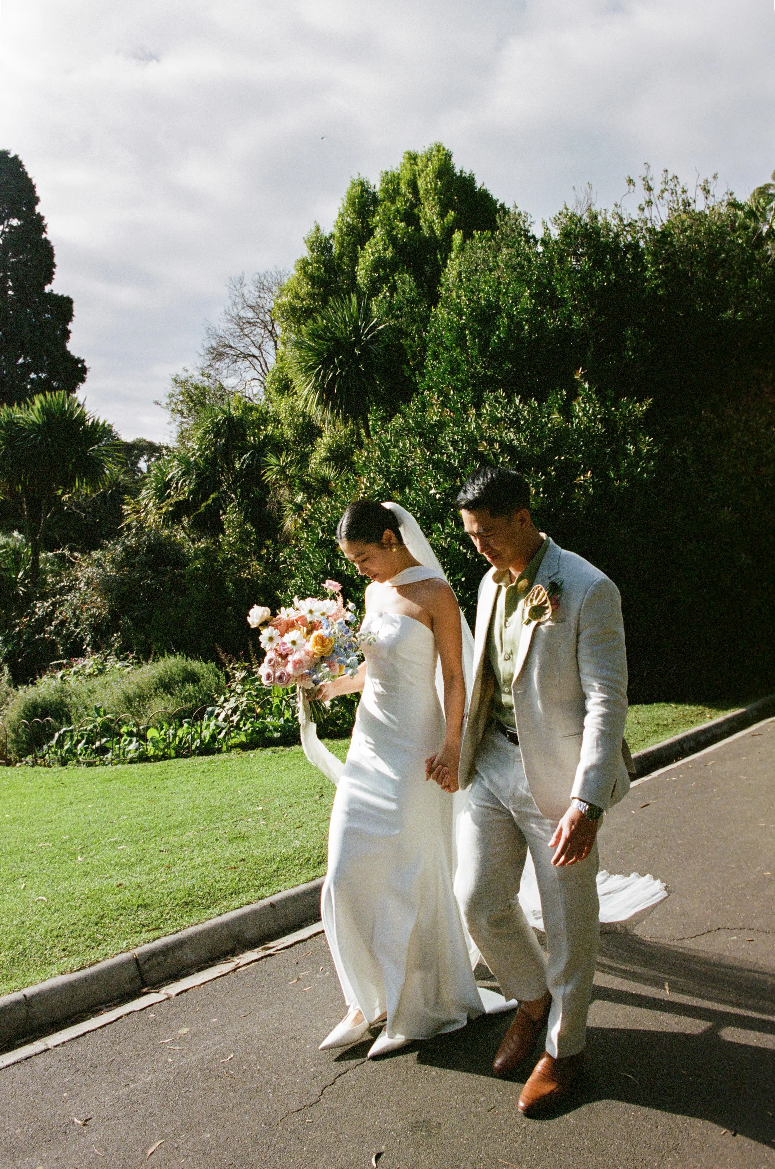 A bride and groom walking hand in hand outdoors on a wedding day, surrounded by green bushes and trees, with cloudy sky overhead.