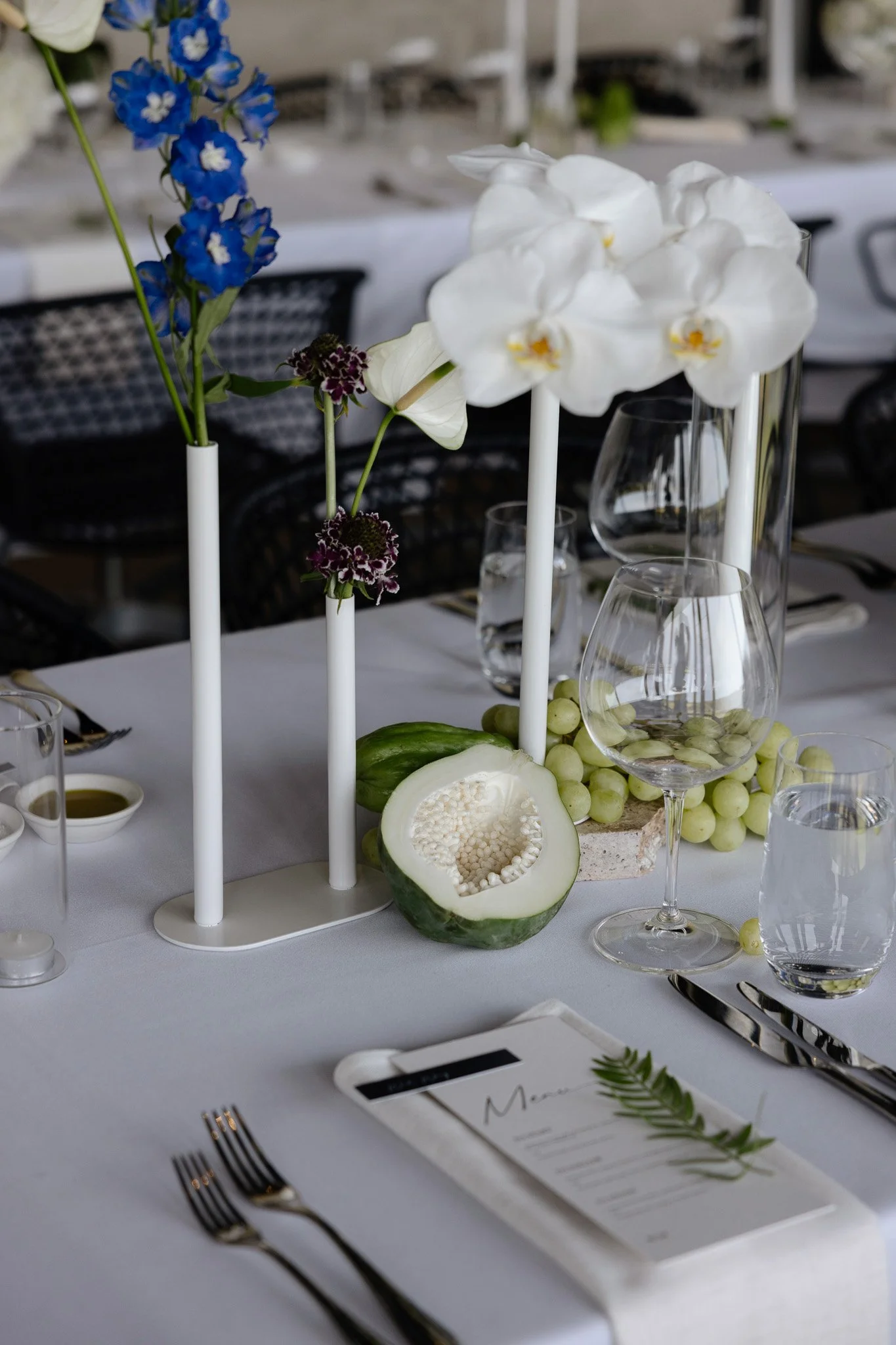 A decorated table set for a formal meal with floral centerpieces, wine glasses, utensils, and a menu. The centerpiece includes white orchids, blue delphiniums, and purple flowers. There is a halved melon with seeds and grapes on the table.
