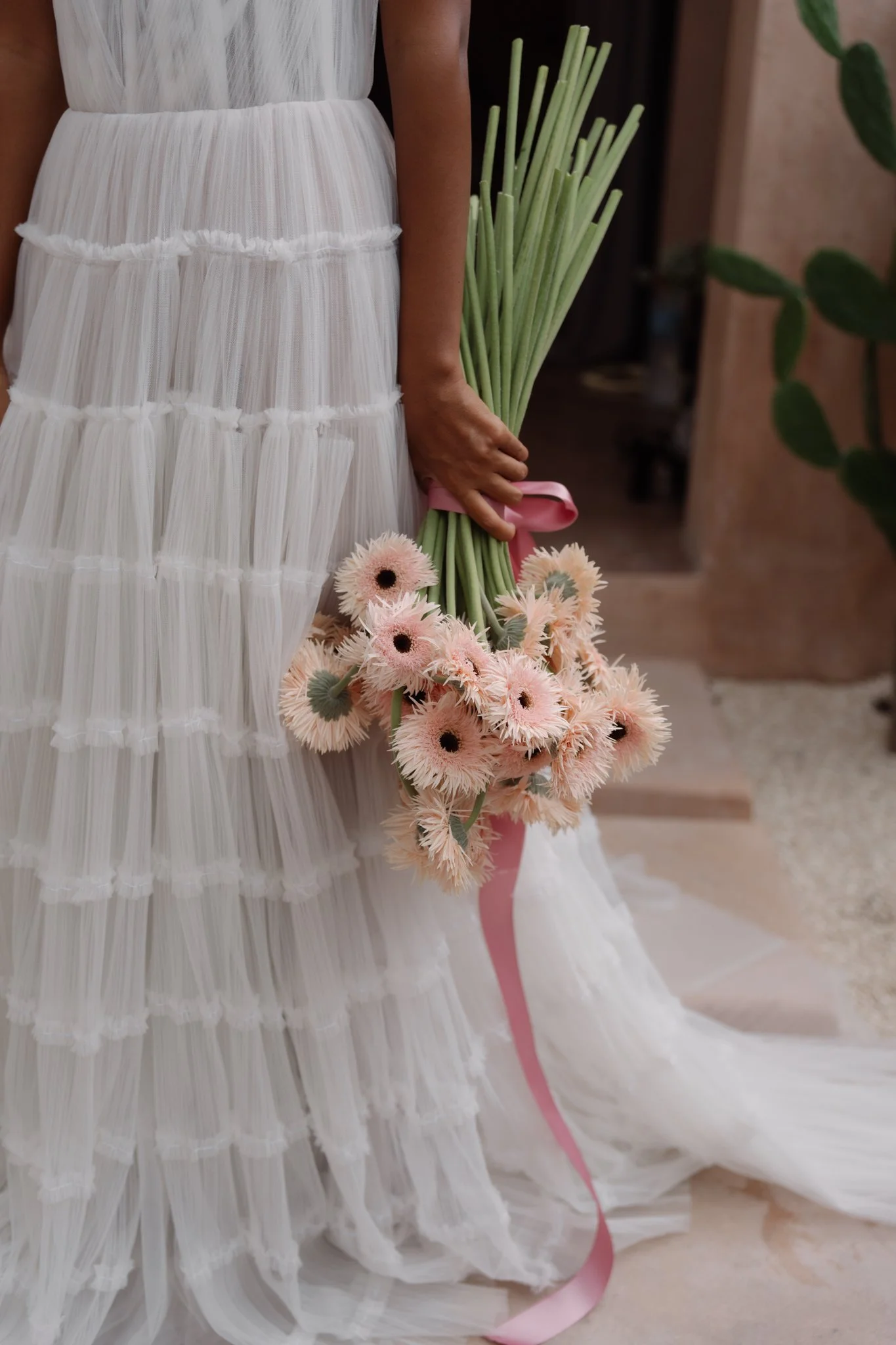 Close-up of a woman holding pink and white flowers, wearing a white dress with layered pleats, indoors with a blurred background and a green plant.