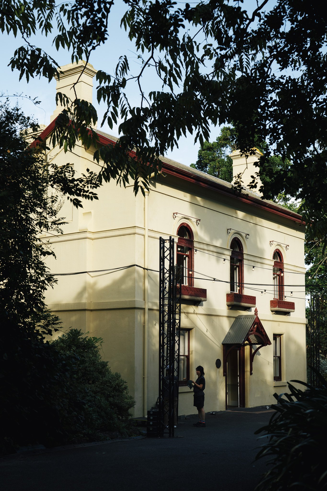 A woman standing outside a cream-colored building with arched windows and a small porch, surrounded by trees and bushes, with a person visible in the background and various equipment or structures in the yard.