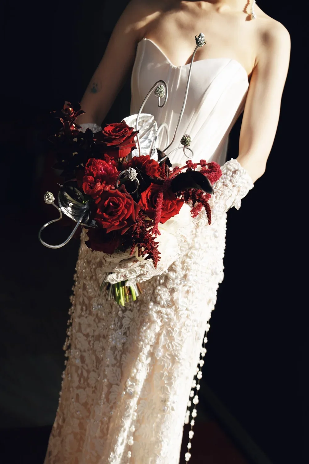 A person in an elegant off-shoulder white dress holds a bouquet of dark red and black flowers with metallic accents, with a dark background.