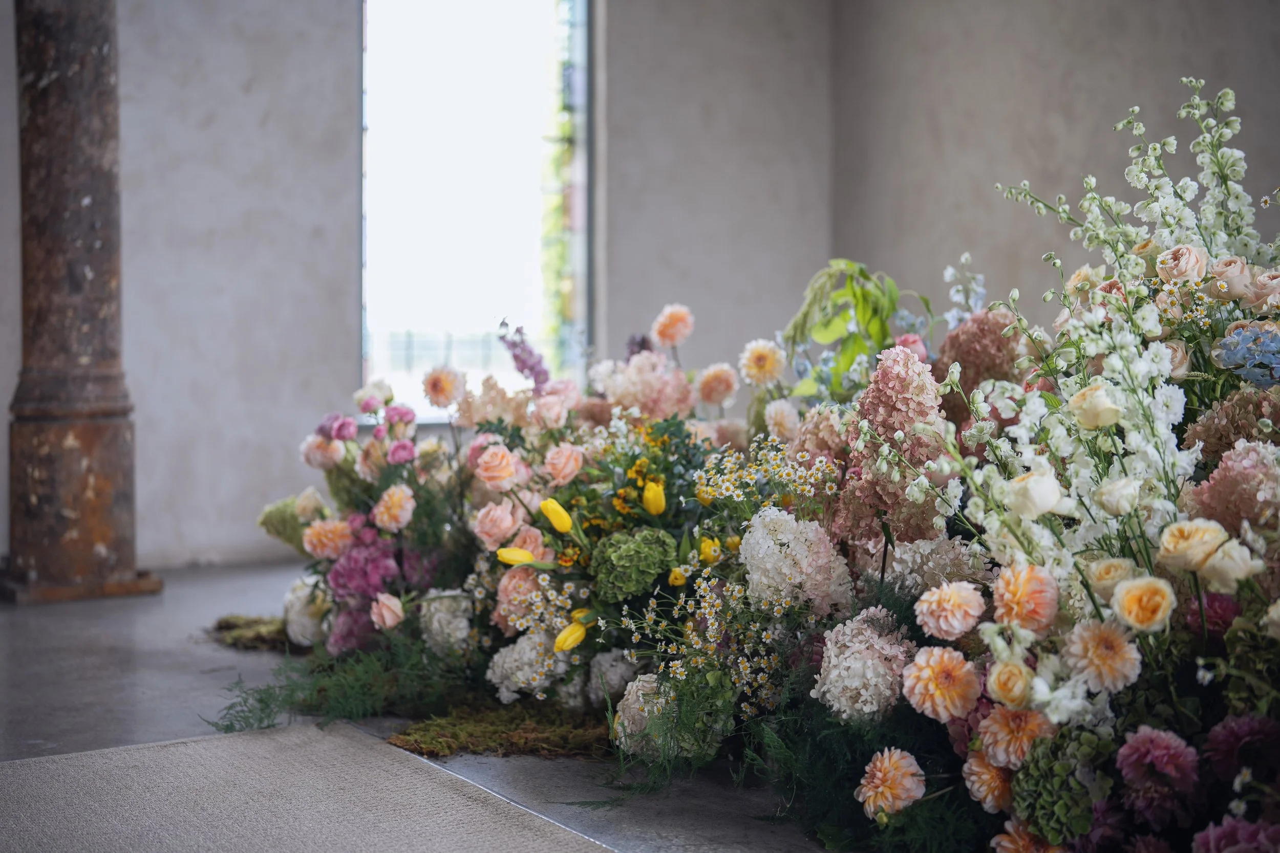 Indoor display of various flowers including roses, hydrangeas, daisies, and others arranged on the floor near a gray concrete wall and window, with a wooden column visible on the left side.