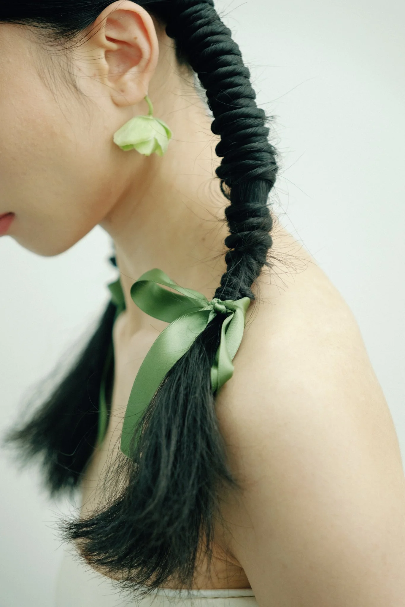 Close-up of a woman's face, ear, and long black braided hair with a green ribbon tied at the end, against a plain white wall background.