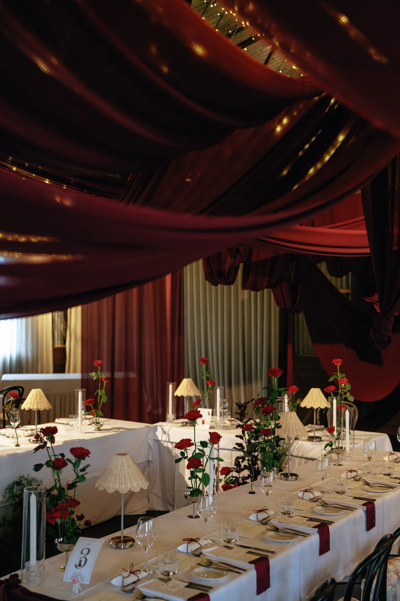 A formal dining setup with multiple tables decorated with white tablecloths, red roses, and beige lampshades. The tables are set with glassware, silverware, and menus or place cards. In the background, there is a dark wooden ceiling with mountain-sha