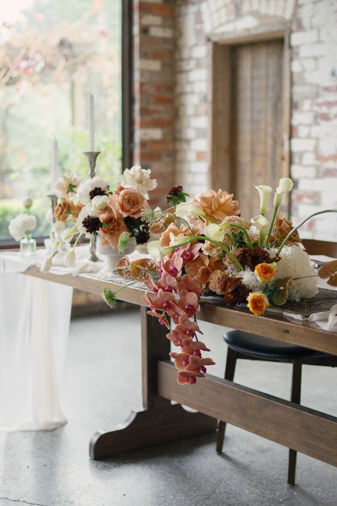 Elegant floral centerpiece with pastel roses, calla lilies, and other flowers on a wooden table in a rustic setting with exposed brick walls and a large window.