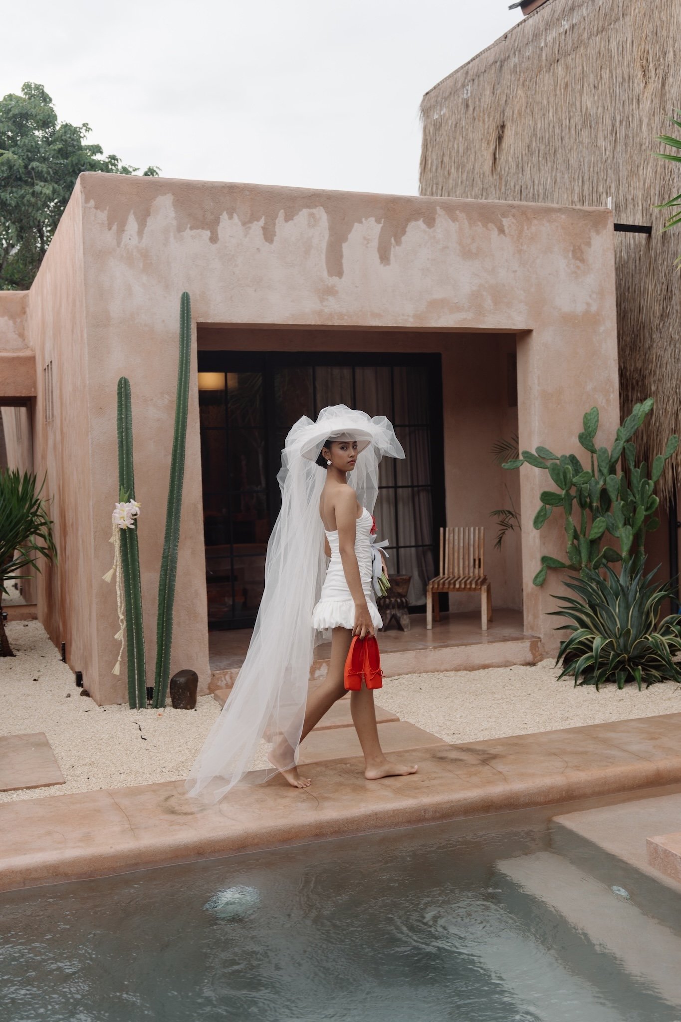 A woman dressed in a white wedding gown with a long train and veil, holding a small red bag, standing barefoot by a poolside with Mediterranean-style pink stucco house, cacti, and lush plants in the background.