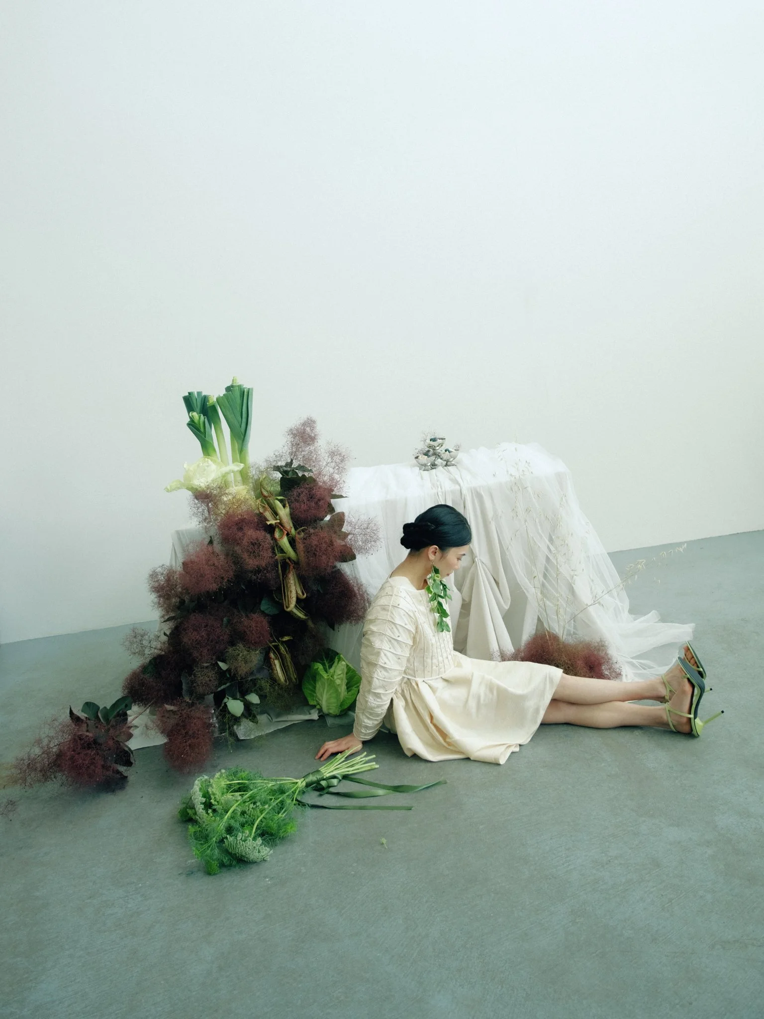 A woman dressed in a beige dress sits on the floor with her legs extended. She is wearing green high heels, has her hair in a bun, and appears to be looking at a smartphone in her hand. Behind her, there is a display of lush flowers and foliage, incl