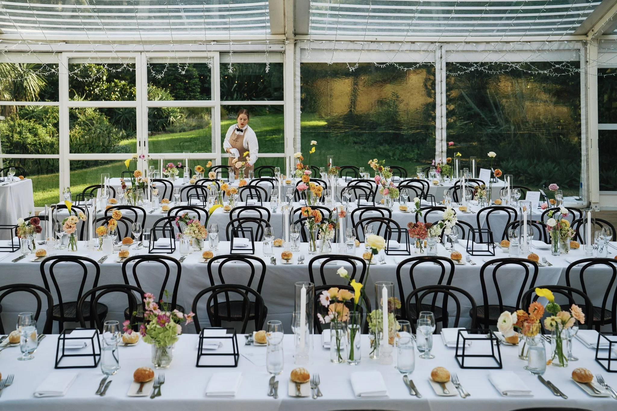 Long banquet tables covered with white tablecloths, decorated with colorful flowers in vases, glassware, bread rolls, and place settings, set inside a greenhouse with large glass windows and string lights, surrounded by greenery.