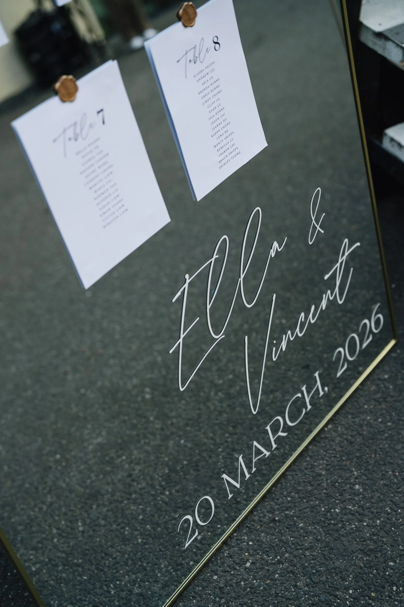 Close-up of a glass sign with white cursive text and a black background, reflecting two printed menus attached with small wood clips, with an asphalt surface underneath.
