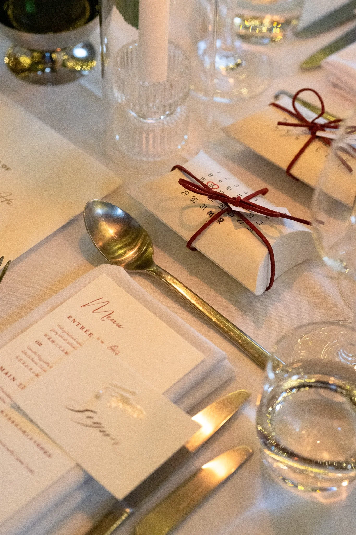 Table setting with menu, wrapped favor boxes tied with ribbon, wine glasses, and a gold-colored spoon.